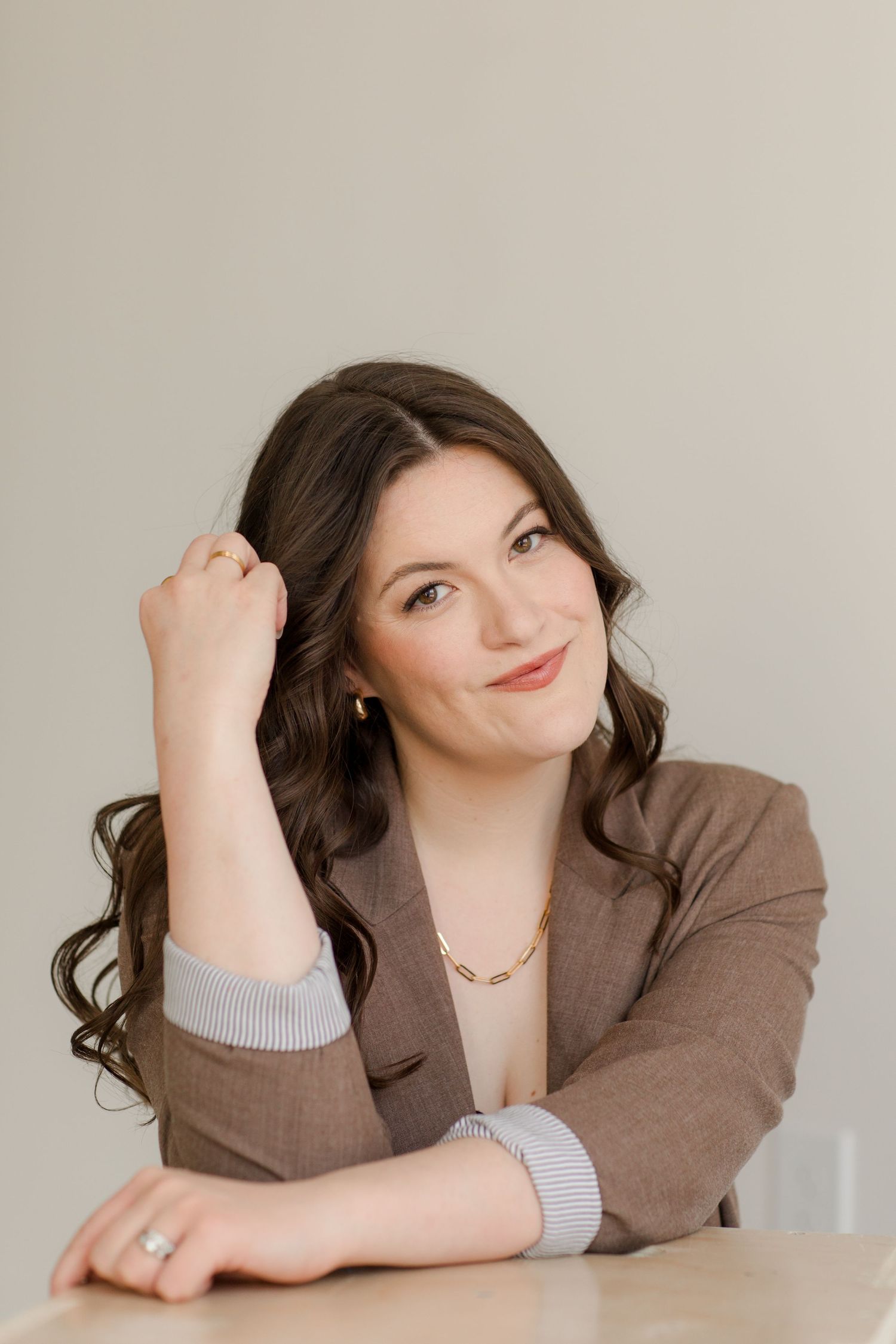 Professional portrait of a person in a brown blazer and gray sweater leaning on a white surface.