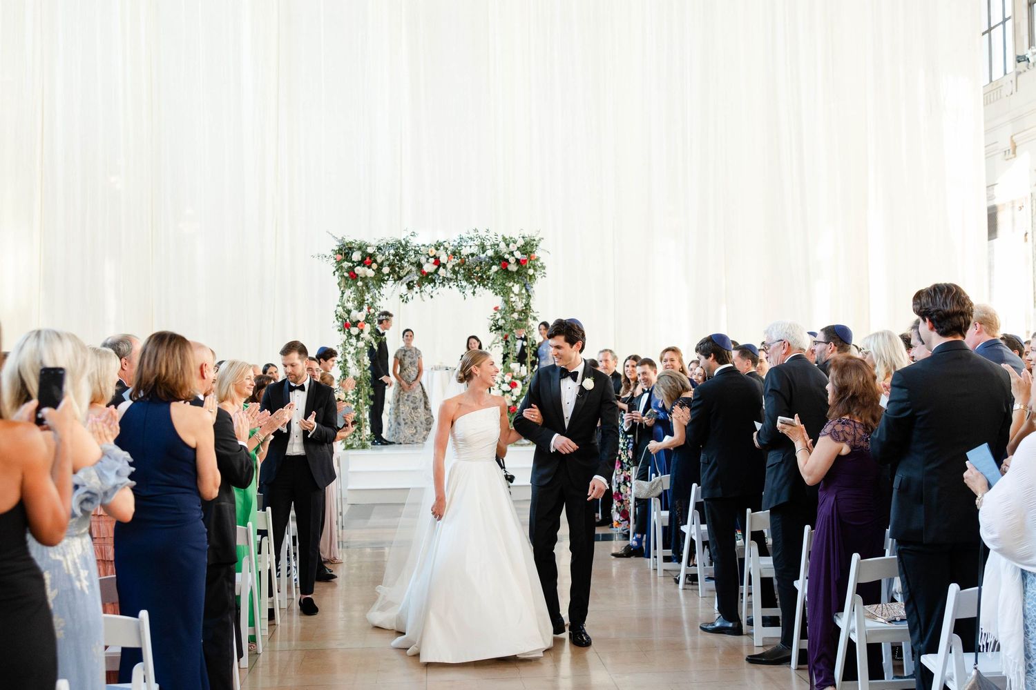 Elegant wedding ceremony in a white industrial venue with floral arch and guests lined along the aisle.