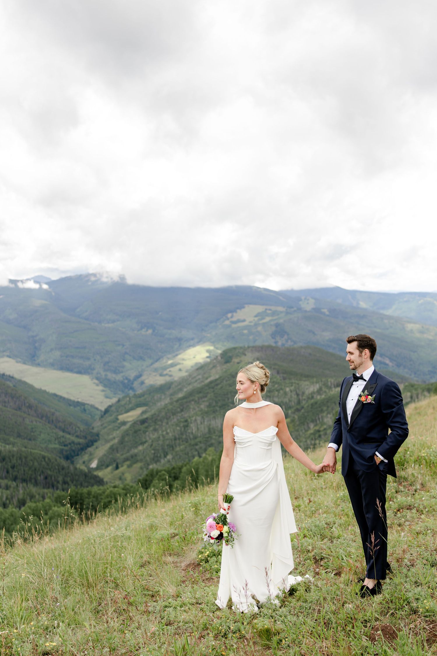 Wedding couple walks hand in hand through mountain meadow with dramatic cloudy sky and evergreen slopes in Vail, Colorado.
