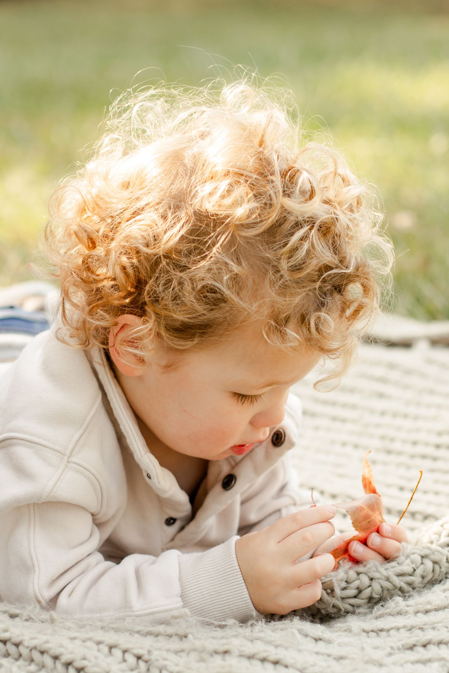 Close-up of a toddler with curly blonde hair examining a toy on a knit blanket.