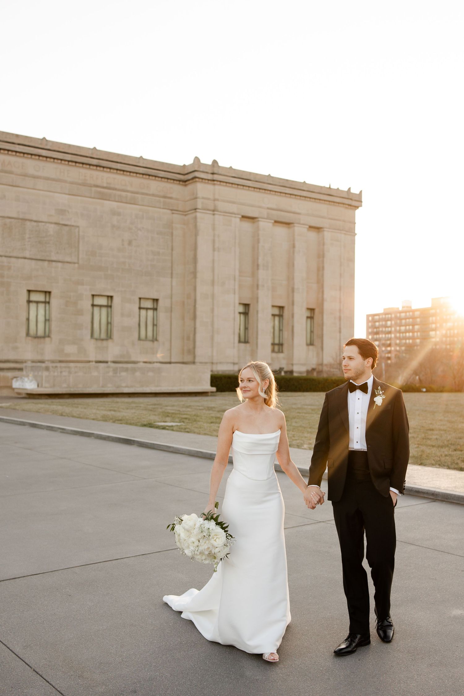 A couple in wedding attire walking outside a grand stone building at sunset at the Nelson Atkins Museum of Art in Kansas City.