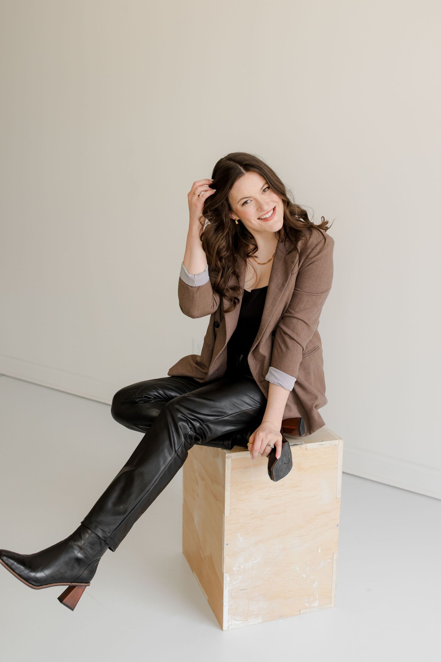 Photographer Hannah Rogg Taylor sits in brown blazer and leather pants sits casually on wooden block against white wall.