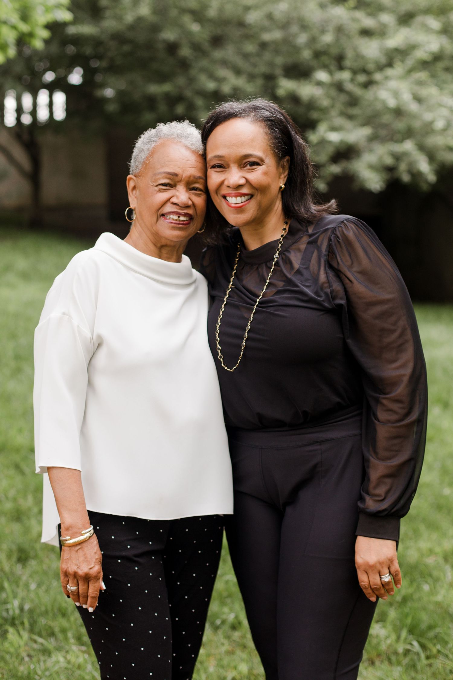 Two people sharing warm embraces and smiles outdoors in a garden setting, wearing contrasting black and white outfits.