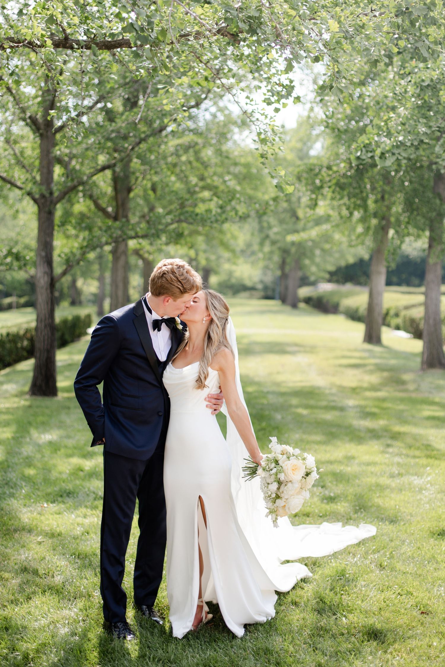 A romantic wedding portrait on a sunlit lawn with trees in the background.