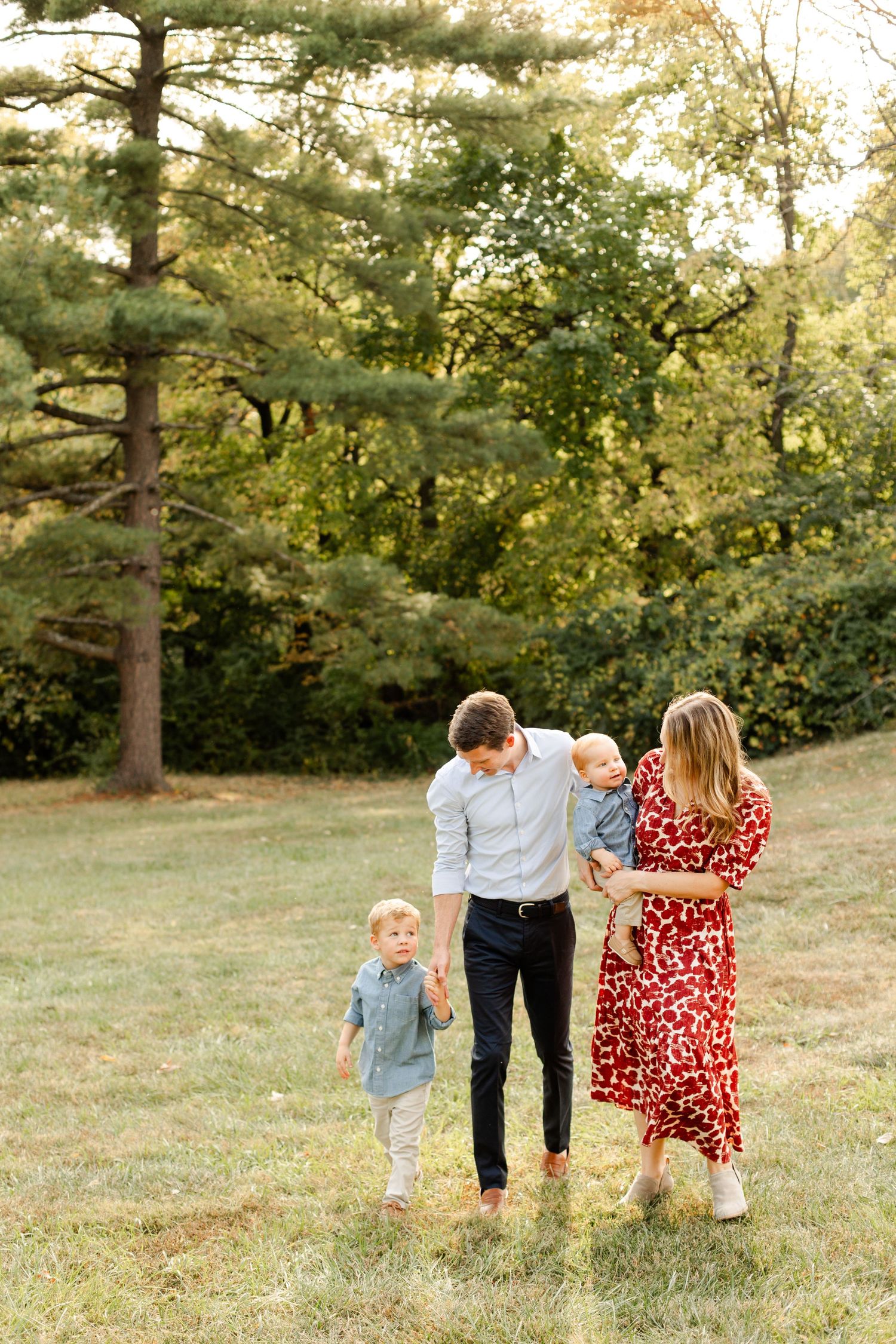 Family walking together on grassy hill with tall pine trees in background. Shawnee Mission Park, Kansas City Family Photos.