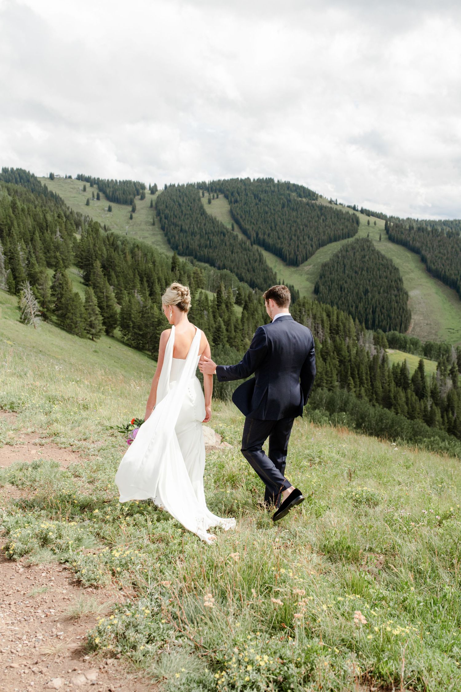 Bride and groom walk hand in hand on a green mountain hillside, with forested ski slopes in the background.