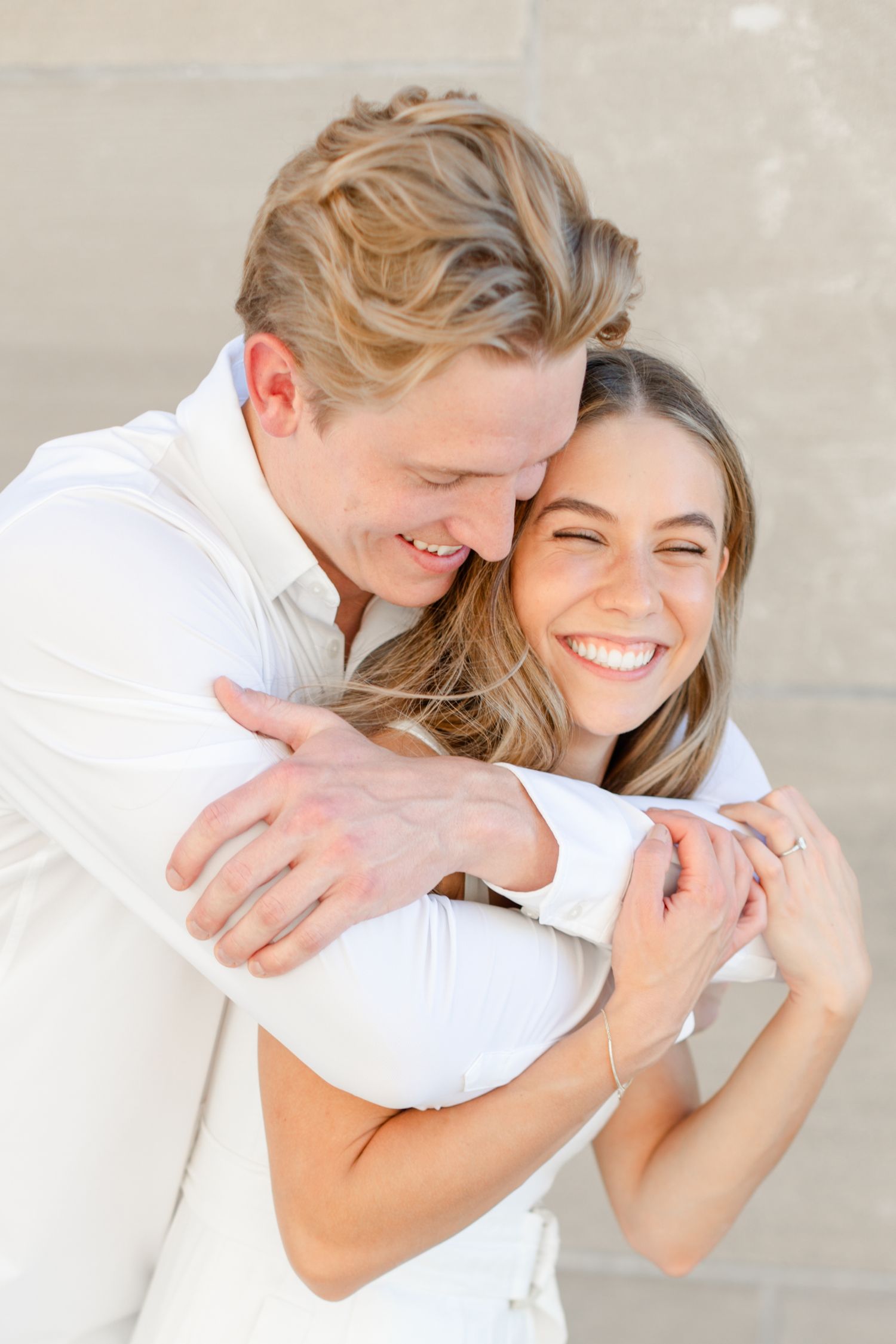 A joyful couple in white embraces against a light colored wall.