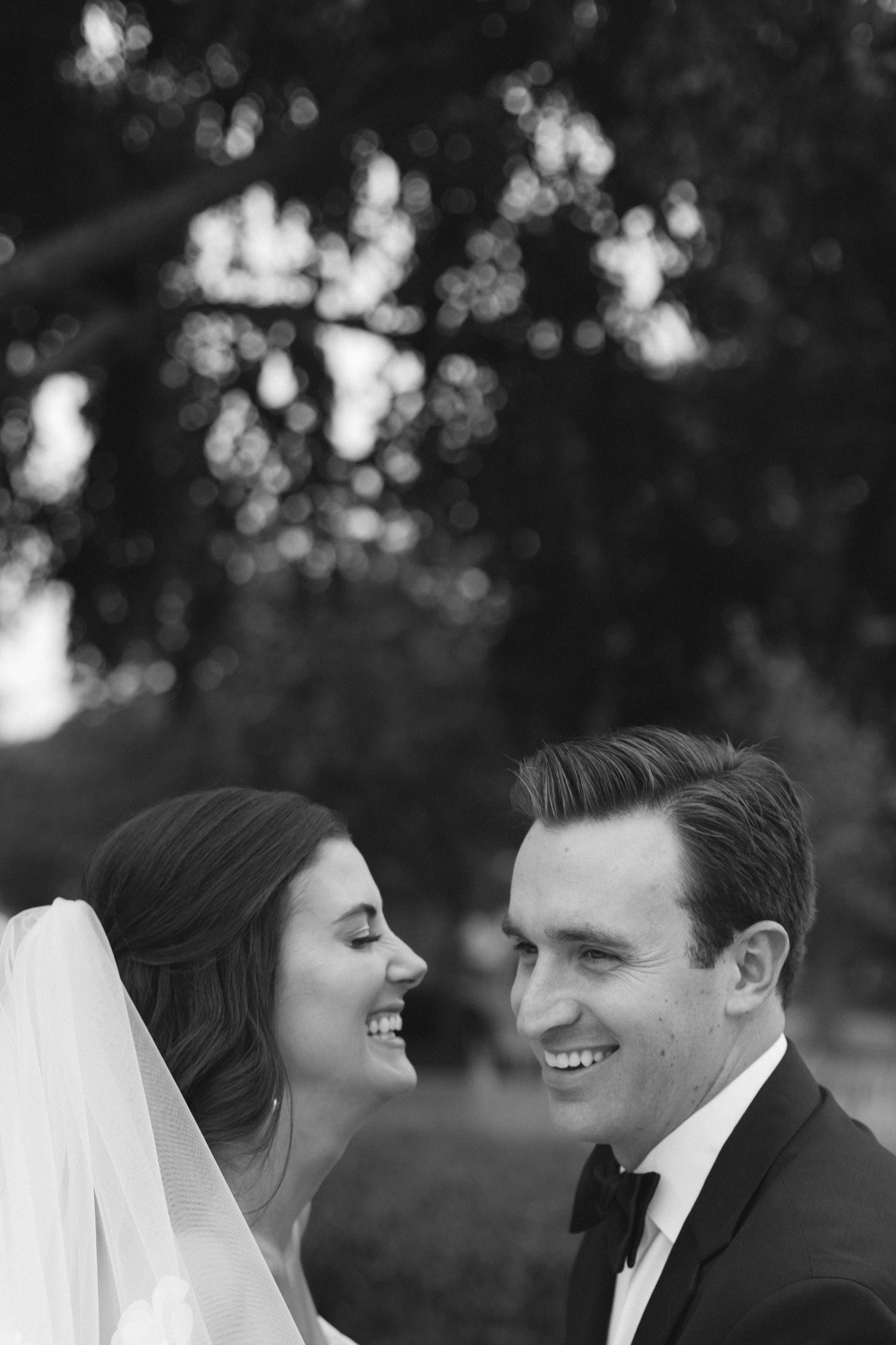 A close up black and white wedding portrait of a happy bride and groom smiling at each other outdoors with bokeh background.