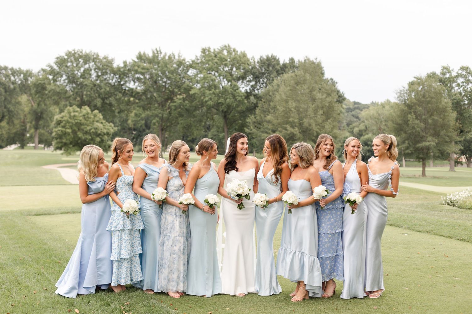 Bridesmaids in flowing light blue dresses stand together with the bride on a lush green golf course.
