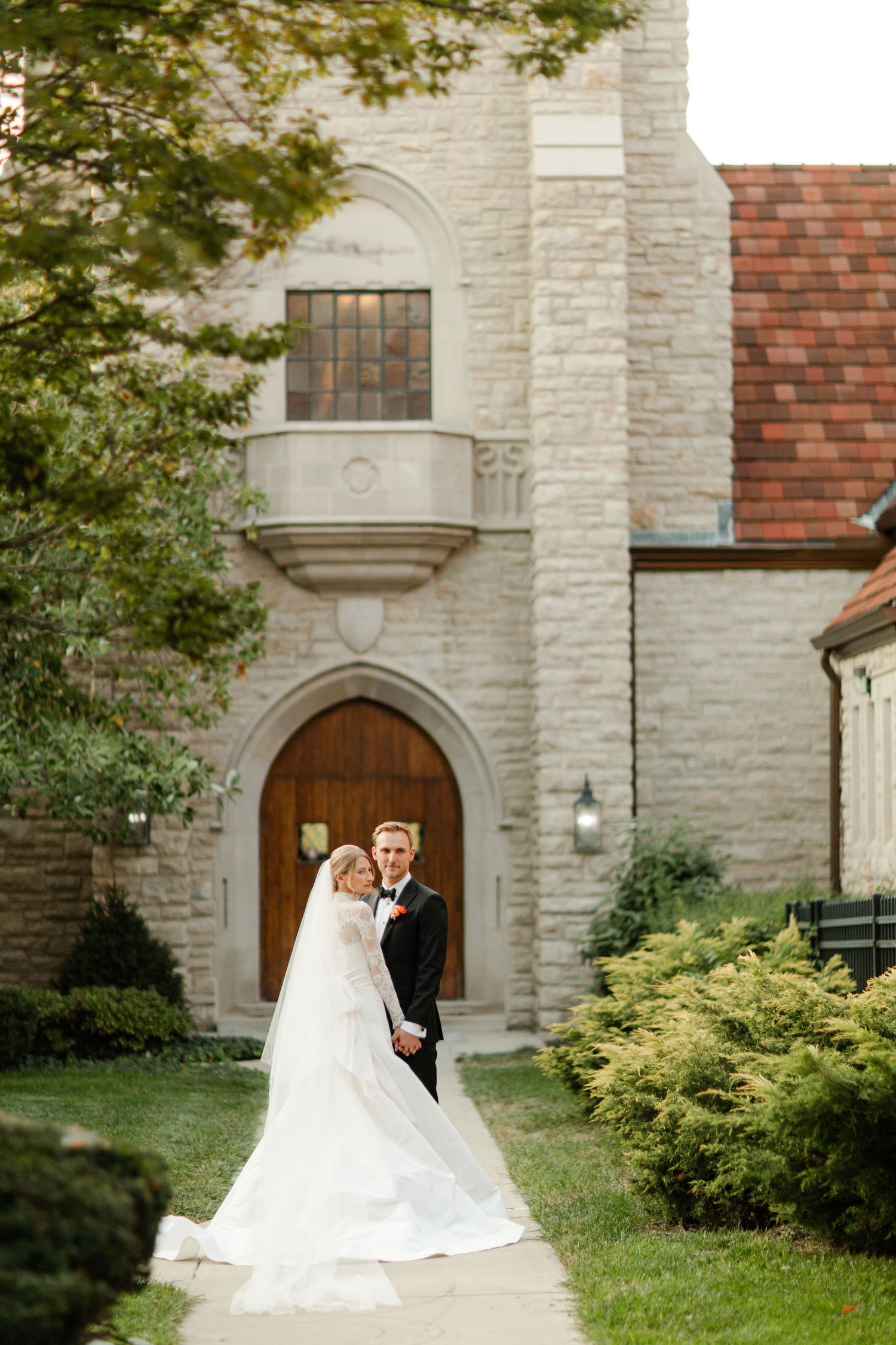 A romantic wedding scene outside a stone church with an arched doorway and red tile roof, surrounded by lush greenery.