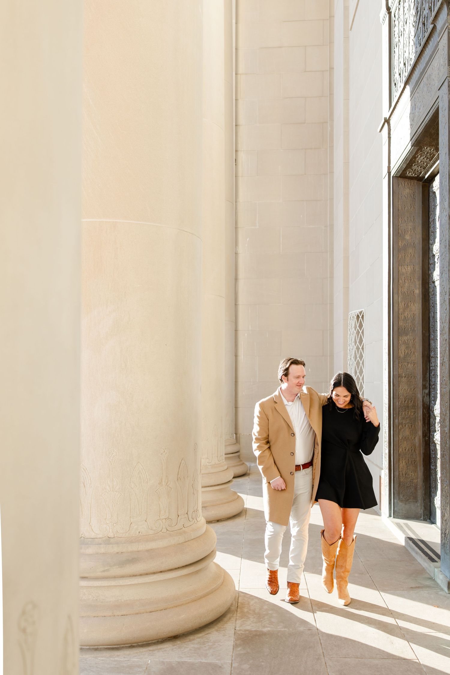 A couple walks together in sunlight between elegant stone columns of a classical building.