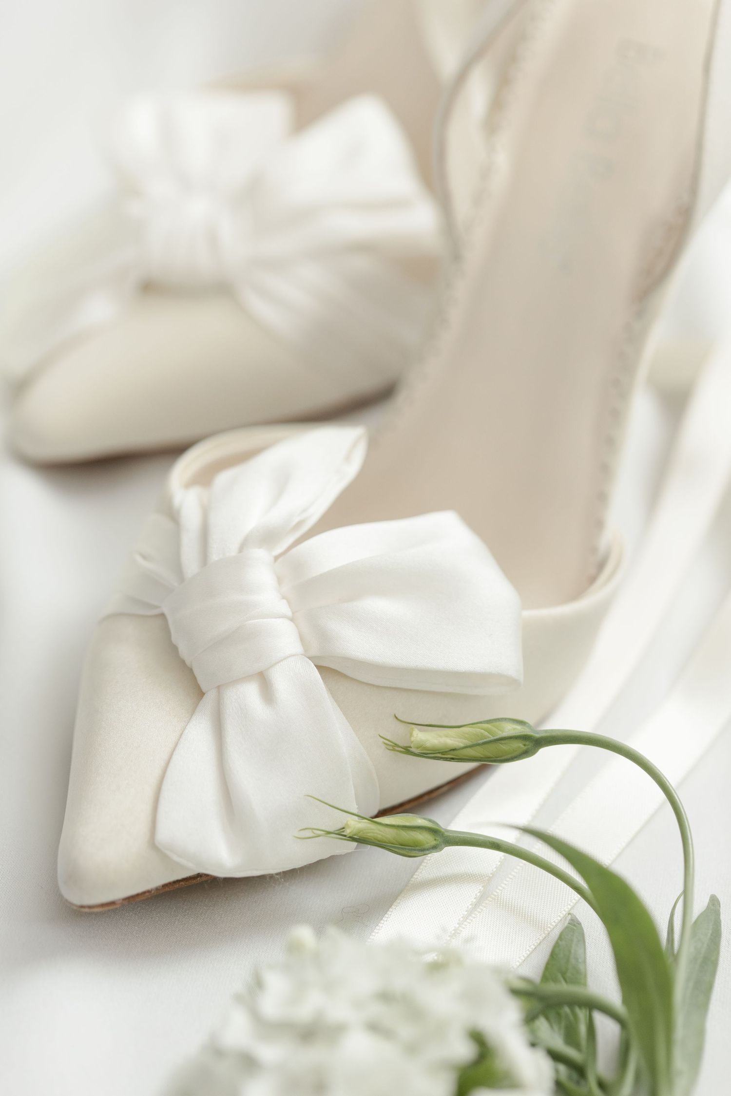 Elegant white bridal shoes with large satin bows and a delicate green flower stem resting on them.