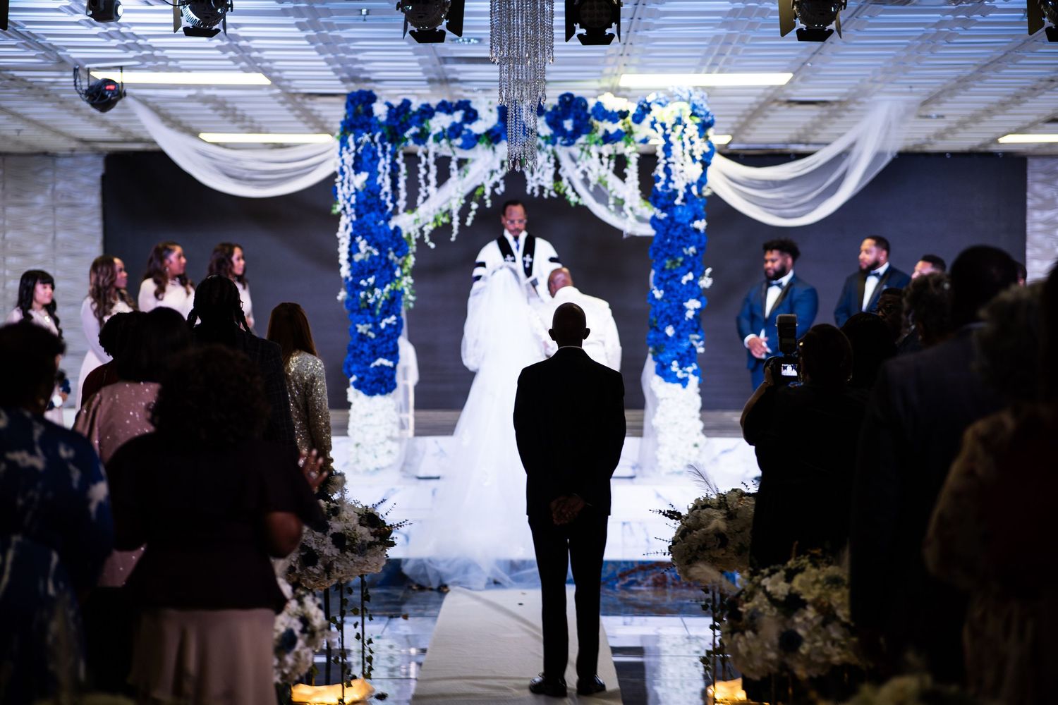 Las Vegas bride and groom join hands as pastor pronounces marriage under blue and white floral stage decor. Groomsmen and bridesmaid stage.