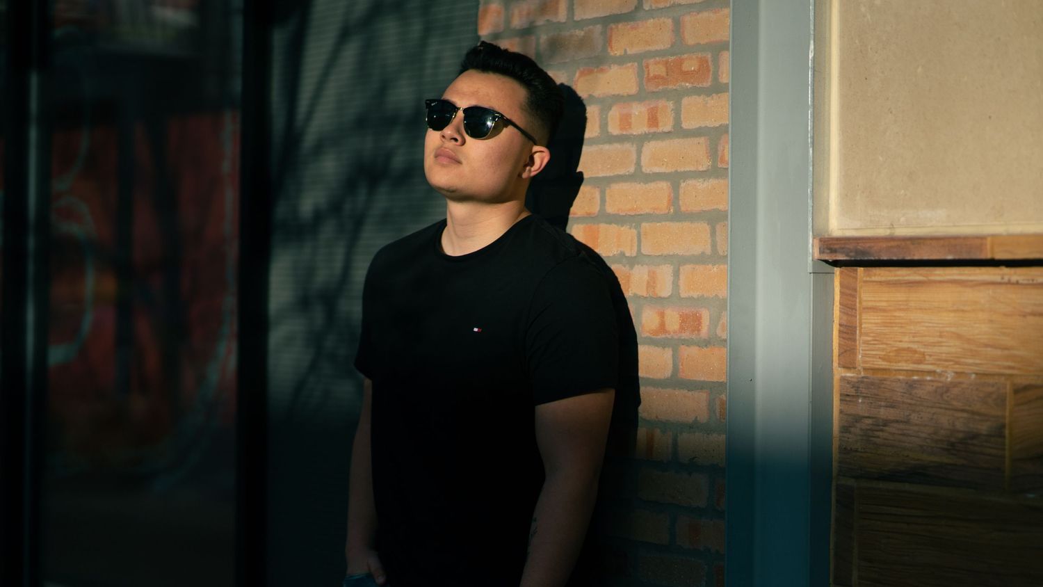 Man in black shirt leaning against brick wall in the shadows.