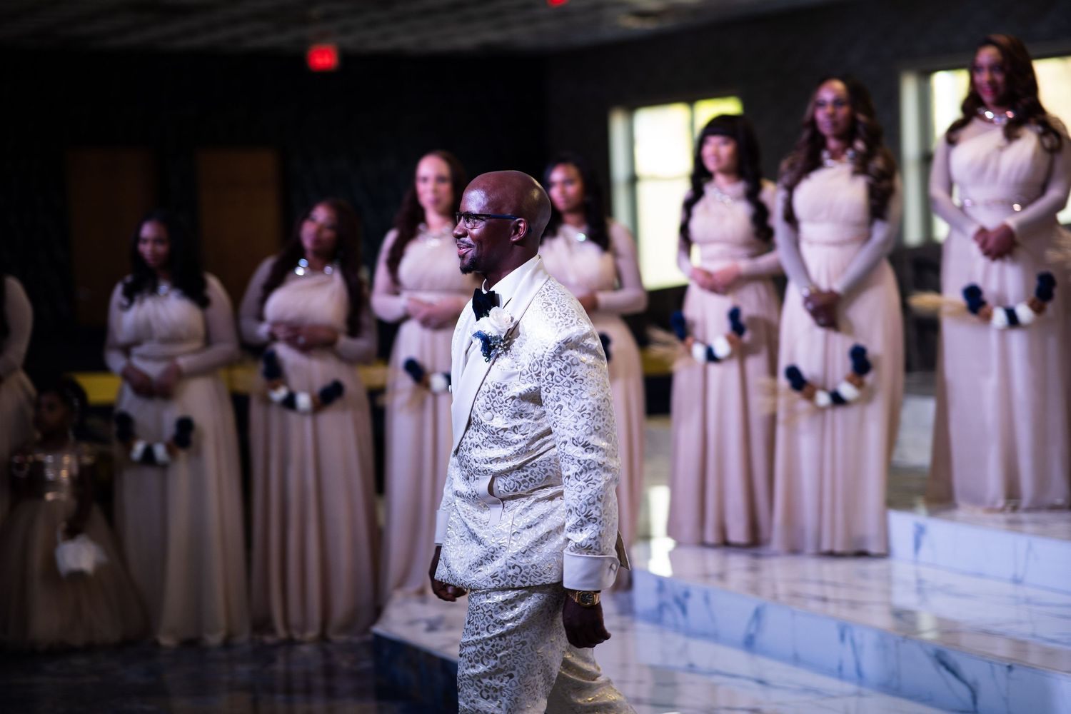 Groom walks to meet bride in front of bridesmaids standing on stairs with floral bouquets.
