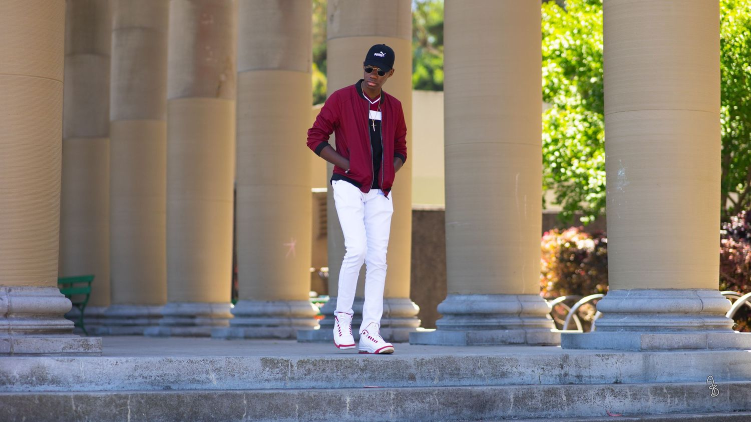 Man in hat and bomber jacket posing against building pillars.