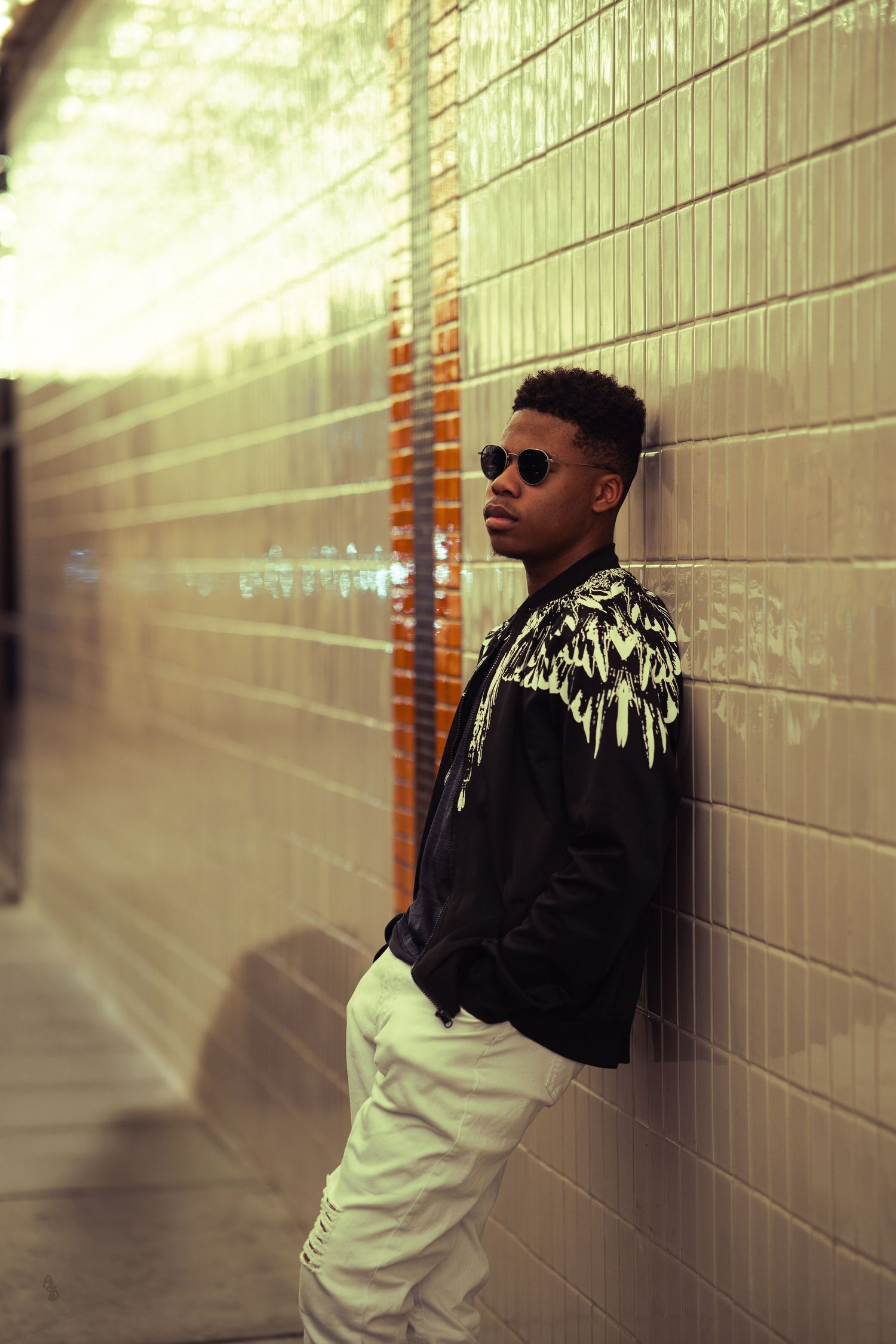 A person in sunglasses leans against a tiled wall in a dimly lit tunnel, wearing a black jacket with a white feather design; a relaxed, cool