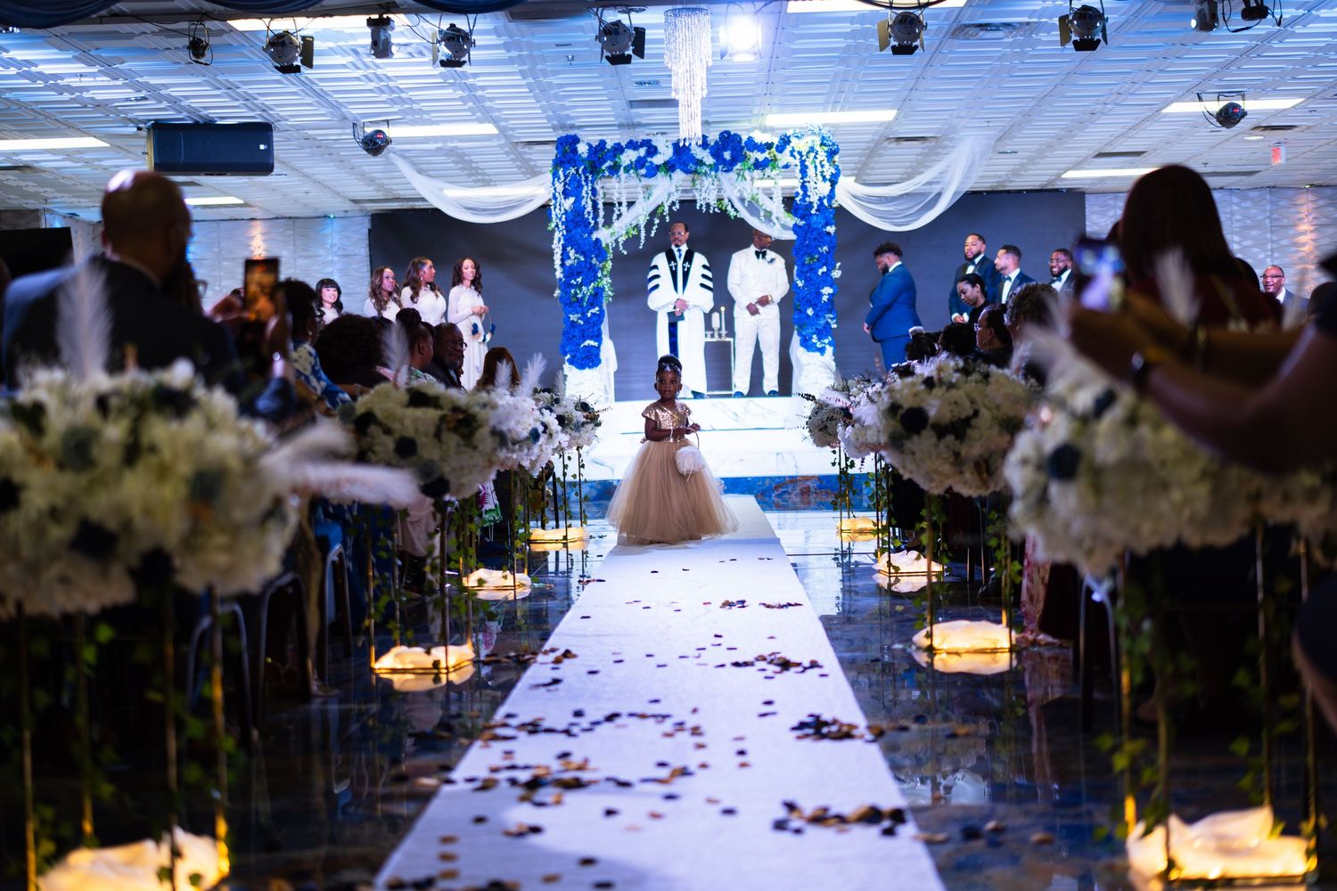 Wedding aisle with flower girl dropping flower petals, Wedding ballroom view. Floral arrangements line aisle.