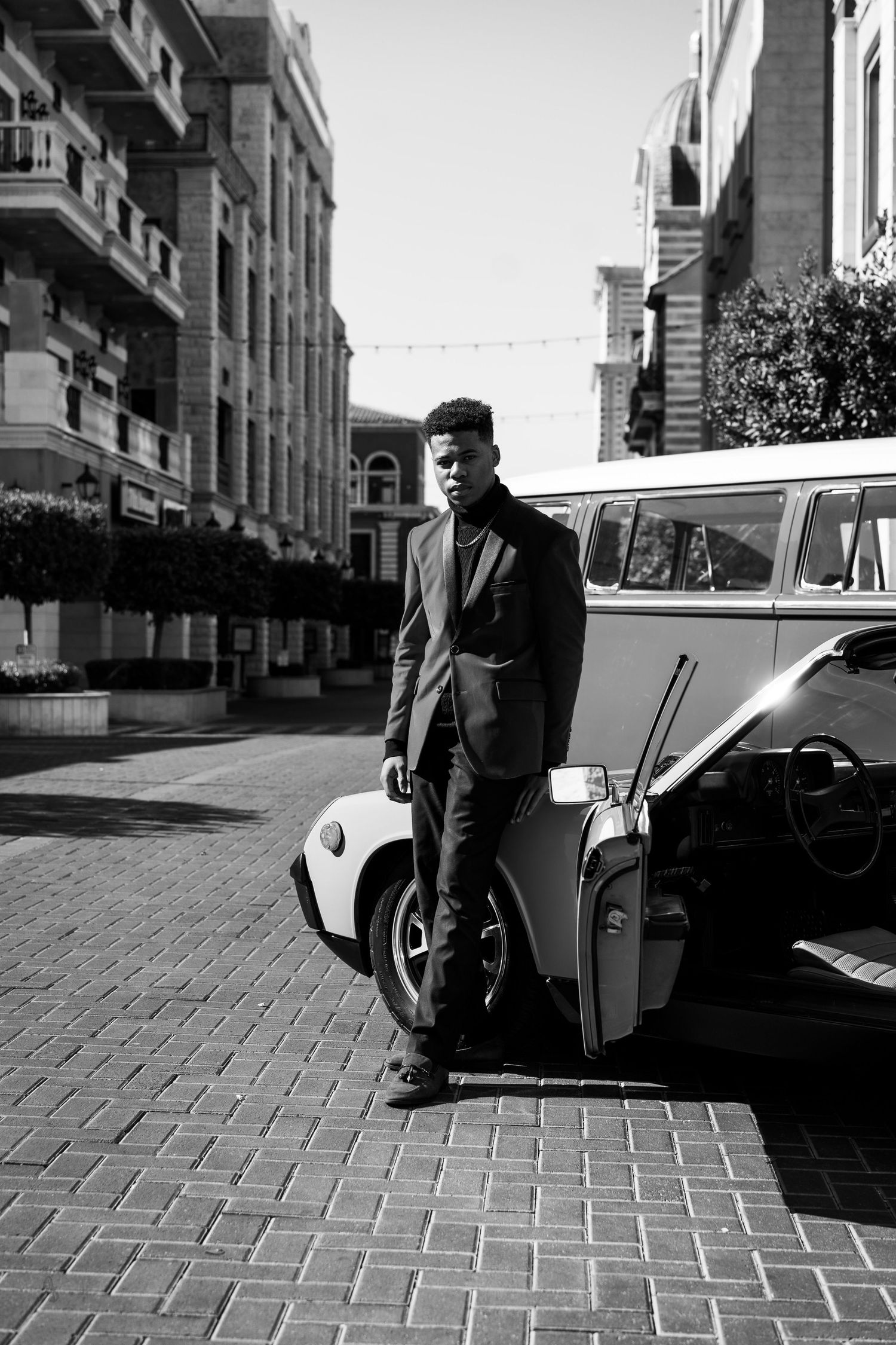 Man posing in front of vintage car and Volkswagen in black and white photo.
