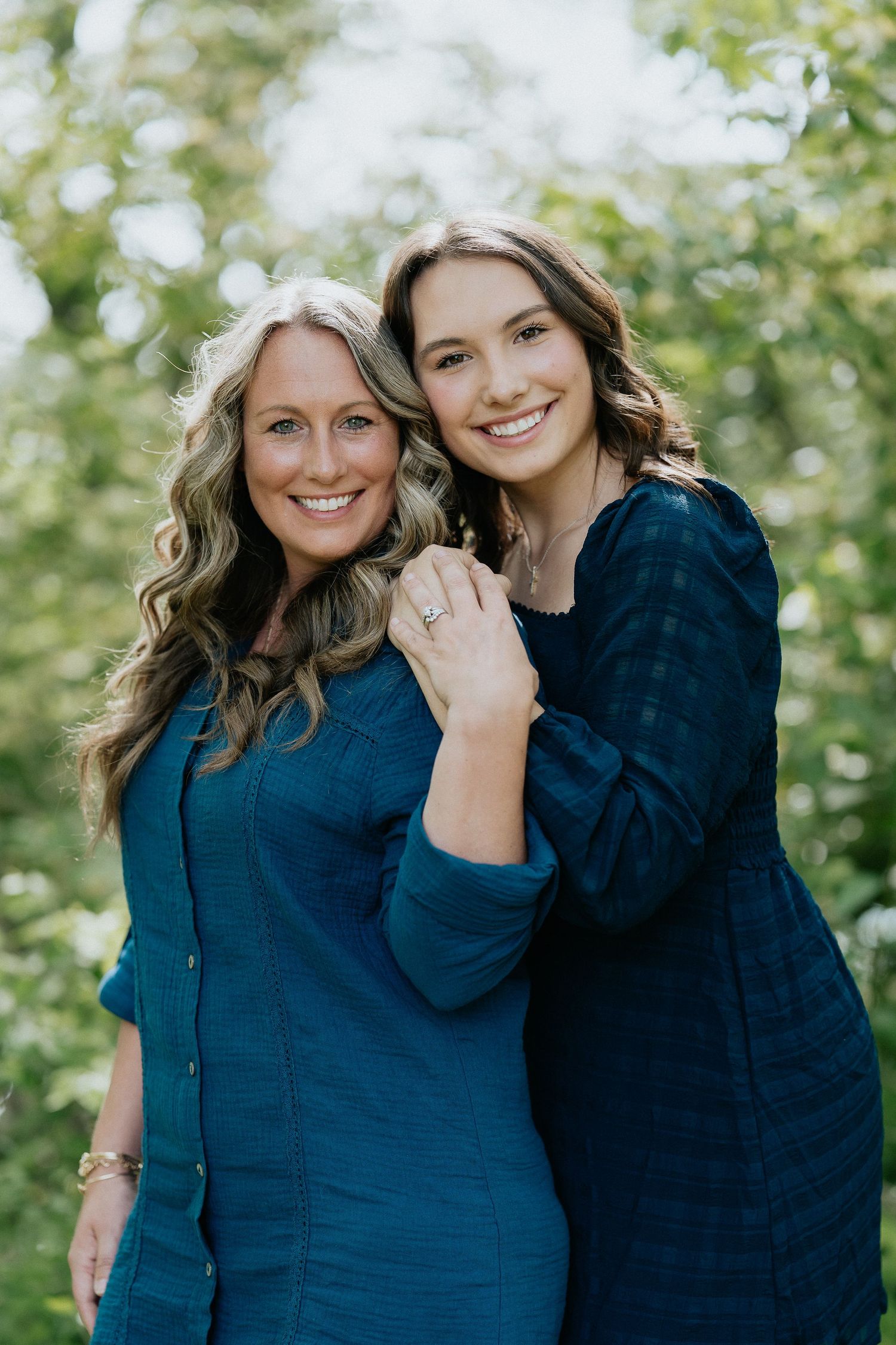 Two people in navy blue sweaters pose together outdoors against a natural green background.