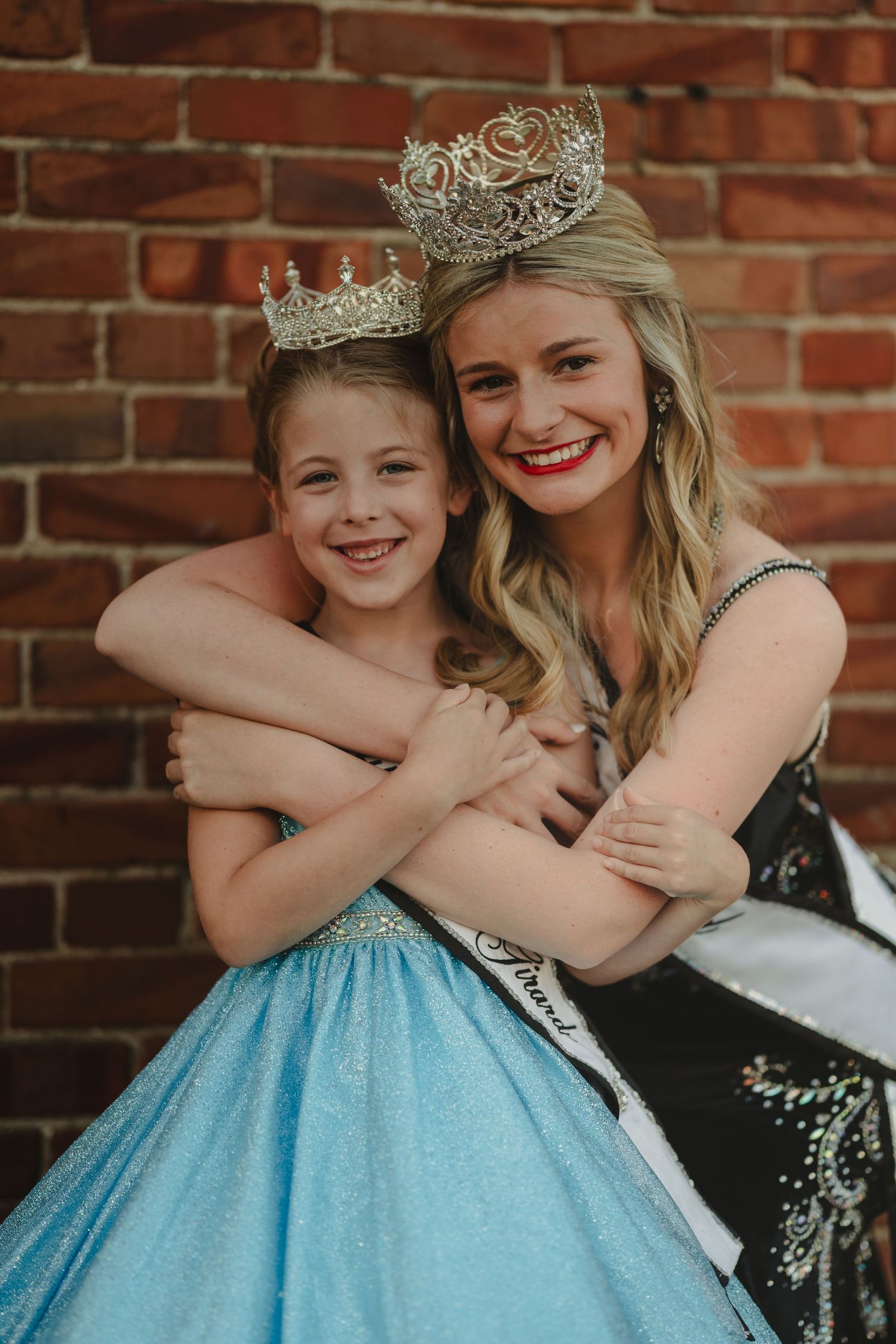 Two girls in formal dresses and crowns embracing against a brick wall background.
