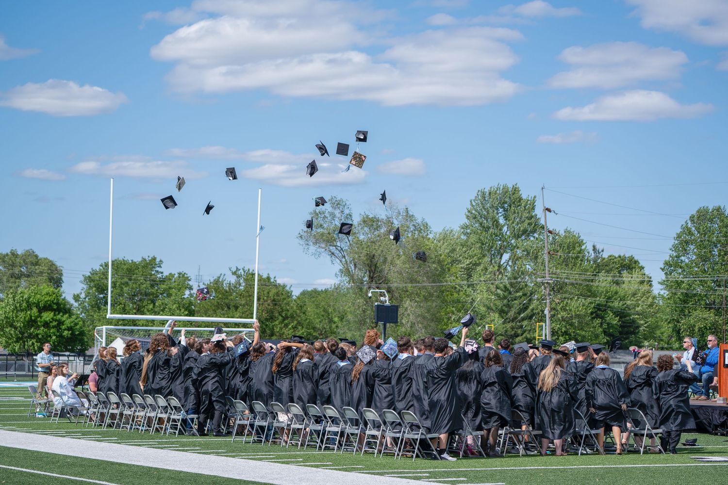 Graduates toss their mortarboards into the blue sky during an outdoor graduation ceremony on a football field.