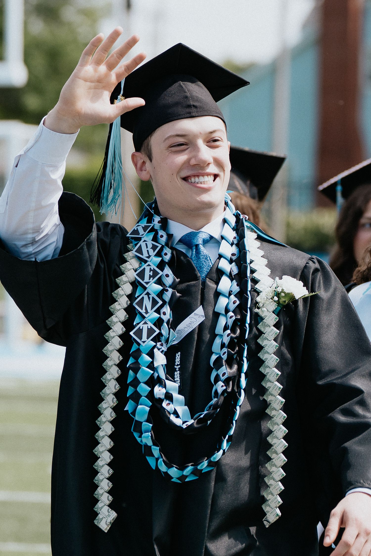 Graduate waving with decorated graduation stole and cap during outdoor ceremony celebration.