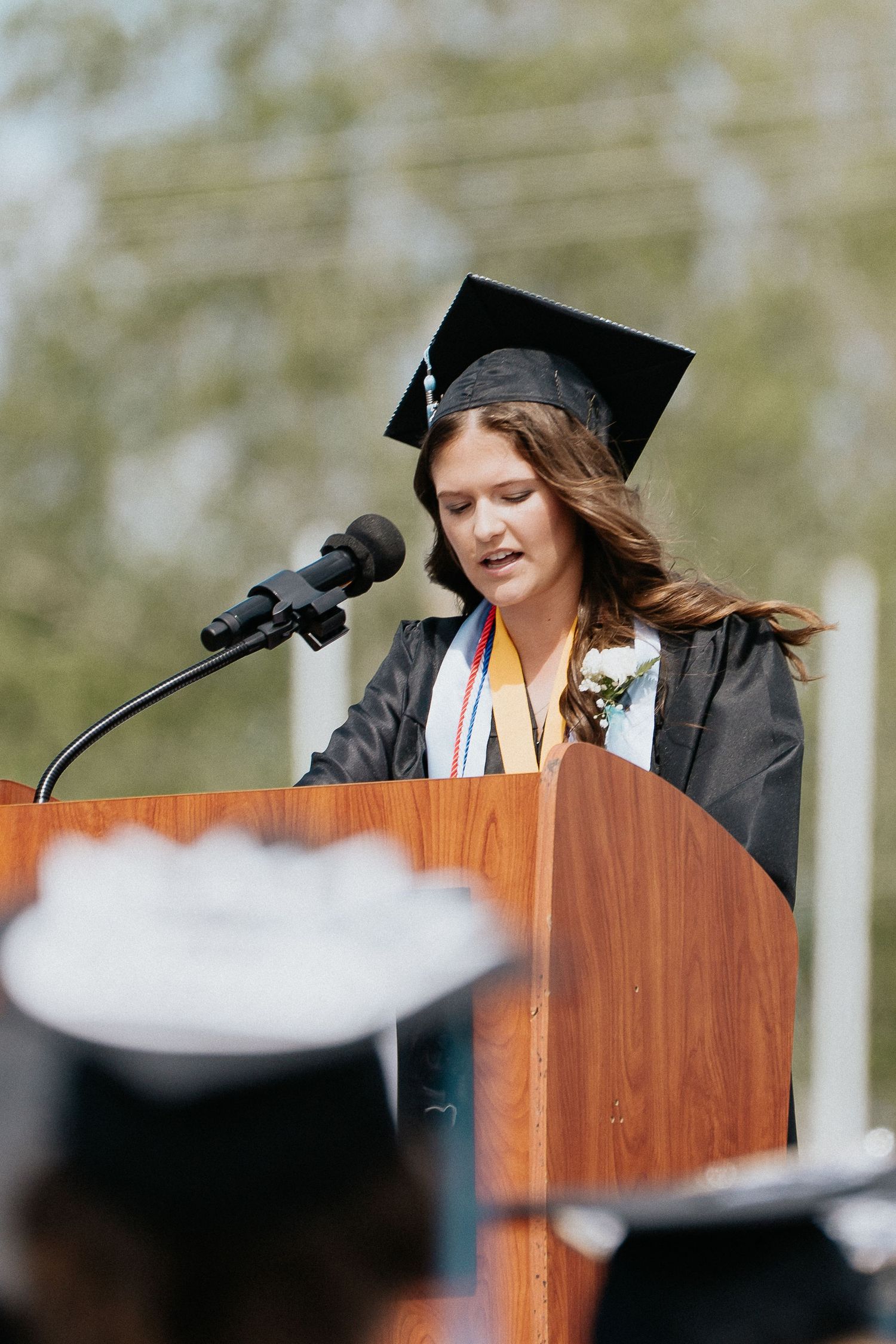 Graduation speaker wearing a black cap and gown stands at a wooden podium delivering a commencement speech outdoors.