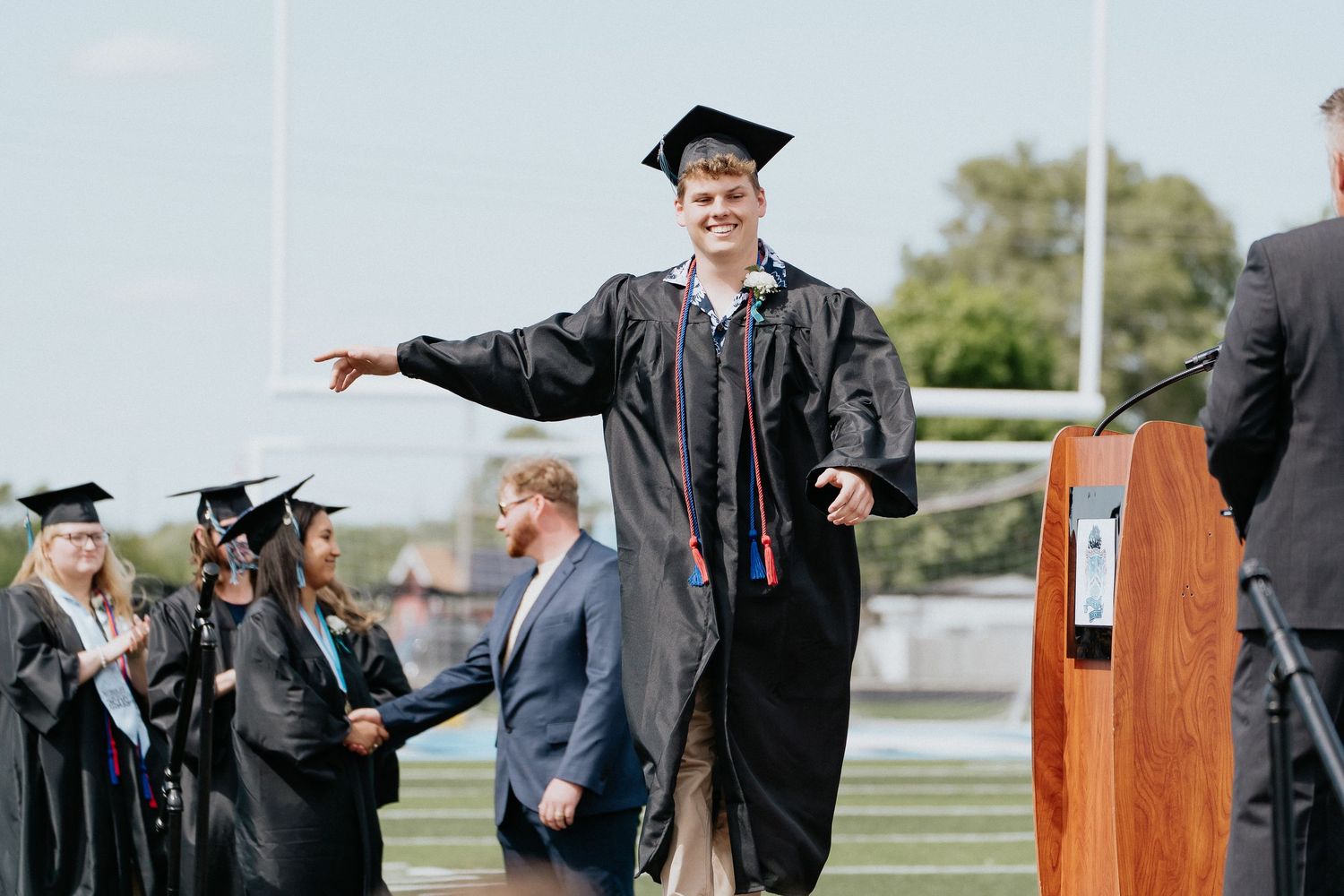 A group of graduates in black caps and gowns celebrates during an outdoor commencement ceremony at a university campus.
