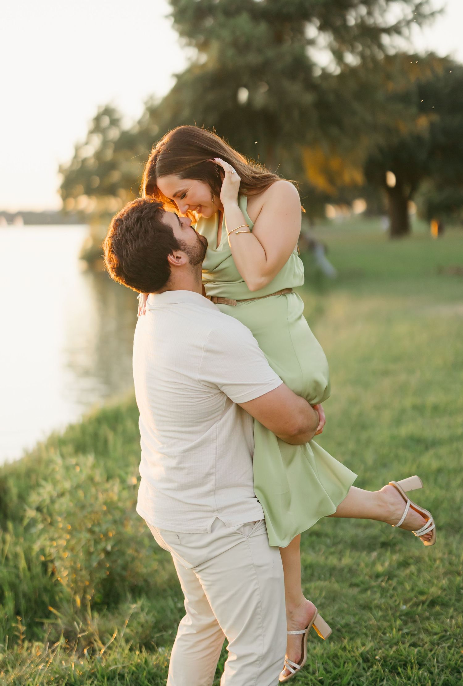 A man lifts a woman in a green dress near a sunlit waterfront, sharing a romantic moment during a golden hour photo session.