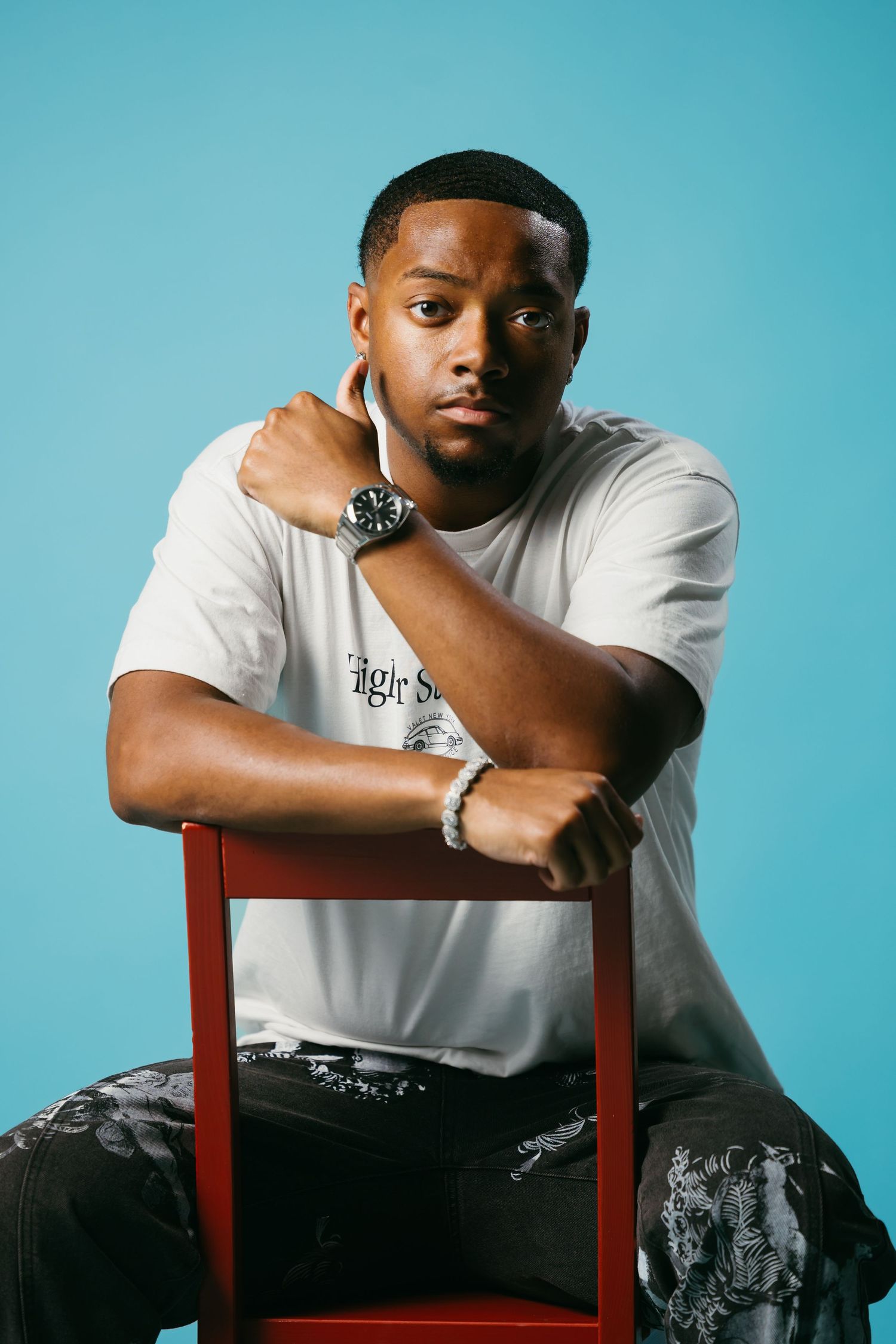 Person in white shirt poses with red chair against blue background wearing watch.