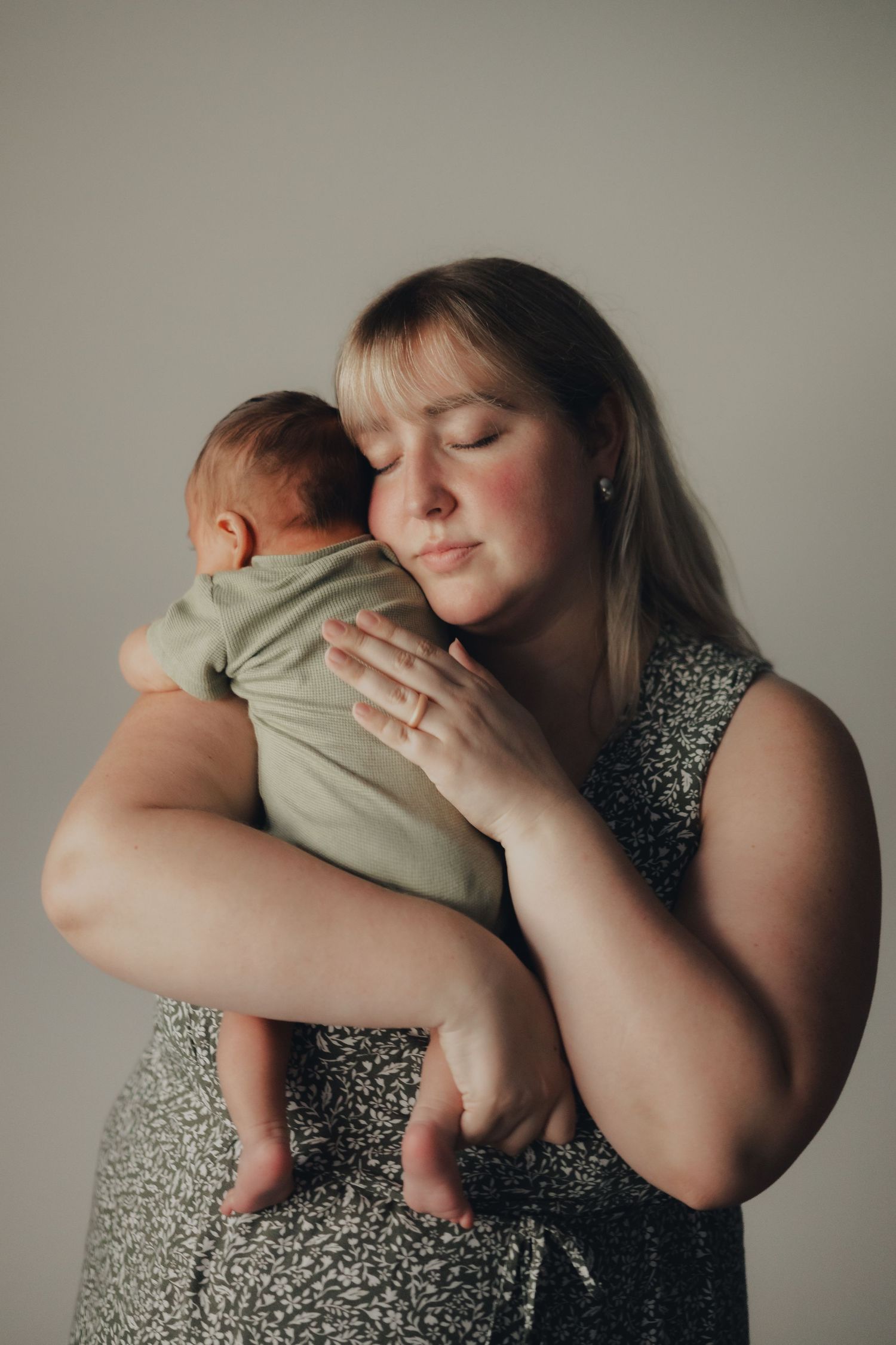 A mother tenderly holds her newborn baby against her cheek with eyes closed in a studio portrait.
