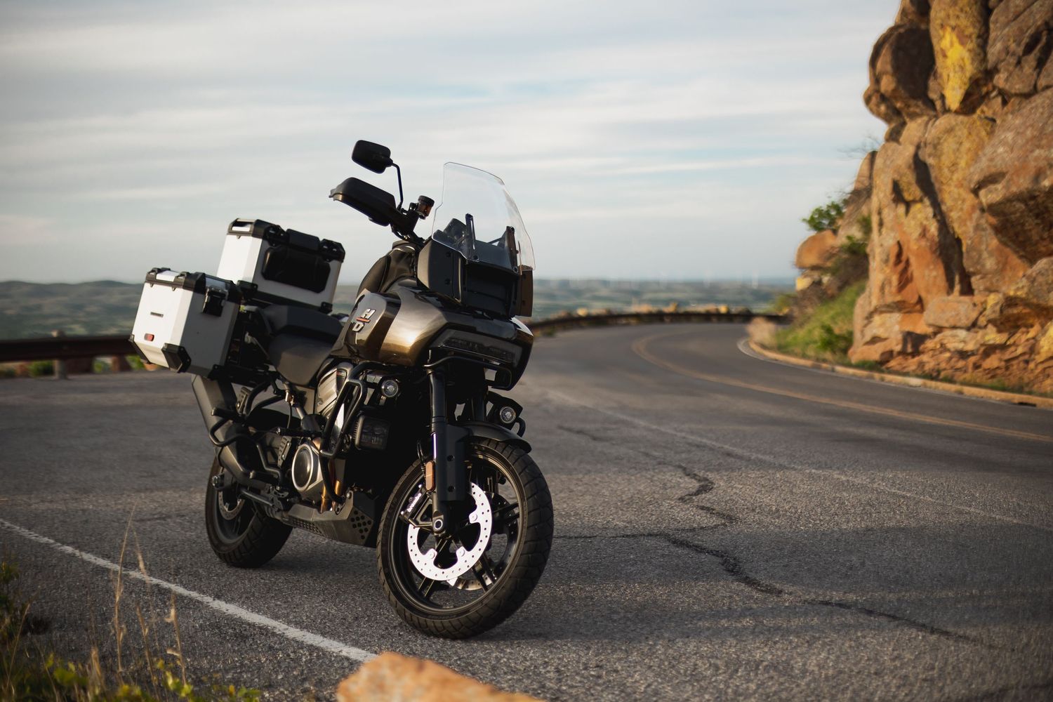 Black adventure motorcycle parked on a winding mountain road with rocky cliffs and scenic sky backdrop.
