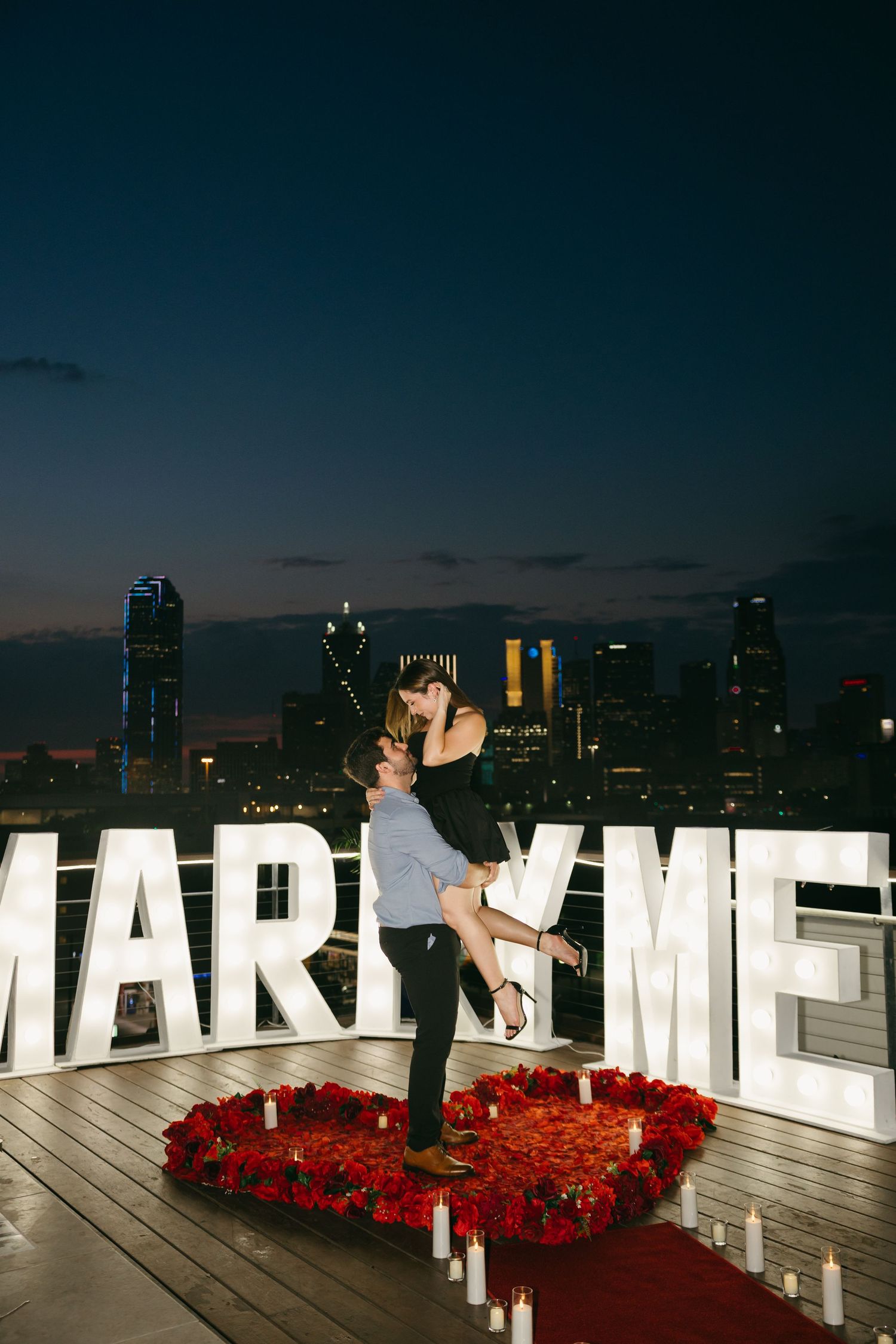 Marriage proposal on rooftop deck with illuminated letters and city skyline at night.