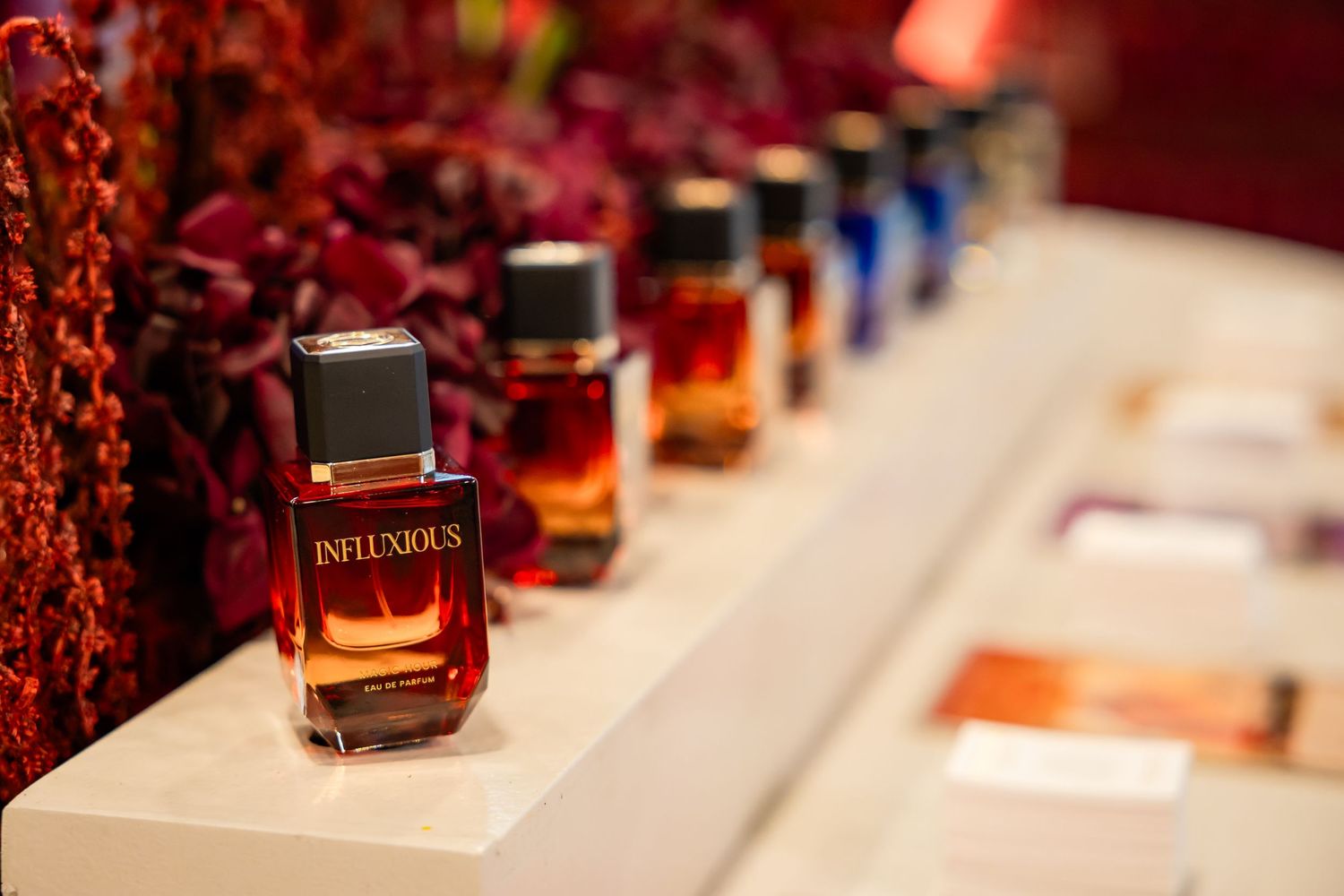 Row of Infuxious luxury perfume bottles displayed on a white shelf with red floral decor in background.