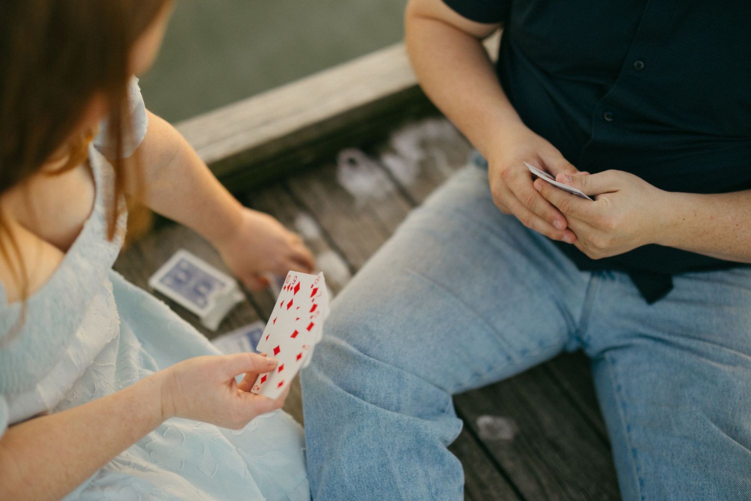 Two people sitting on wooden steps playing cards together, holding a hand of playing cards outdoors.