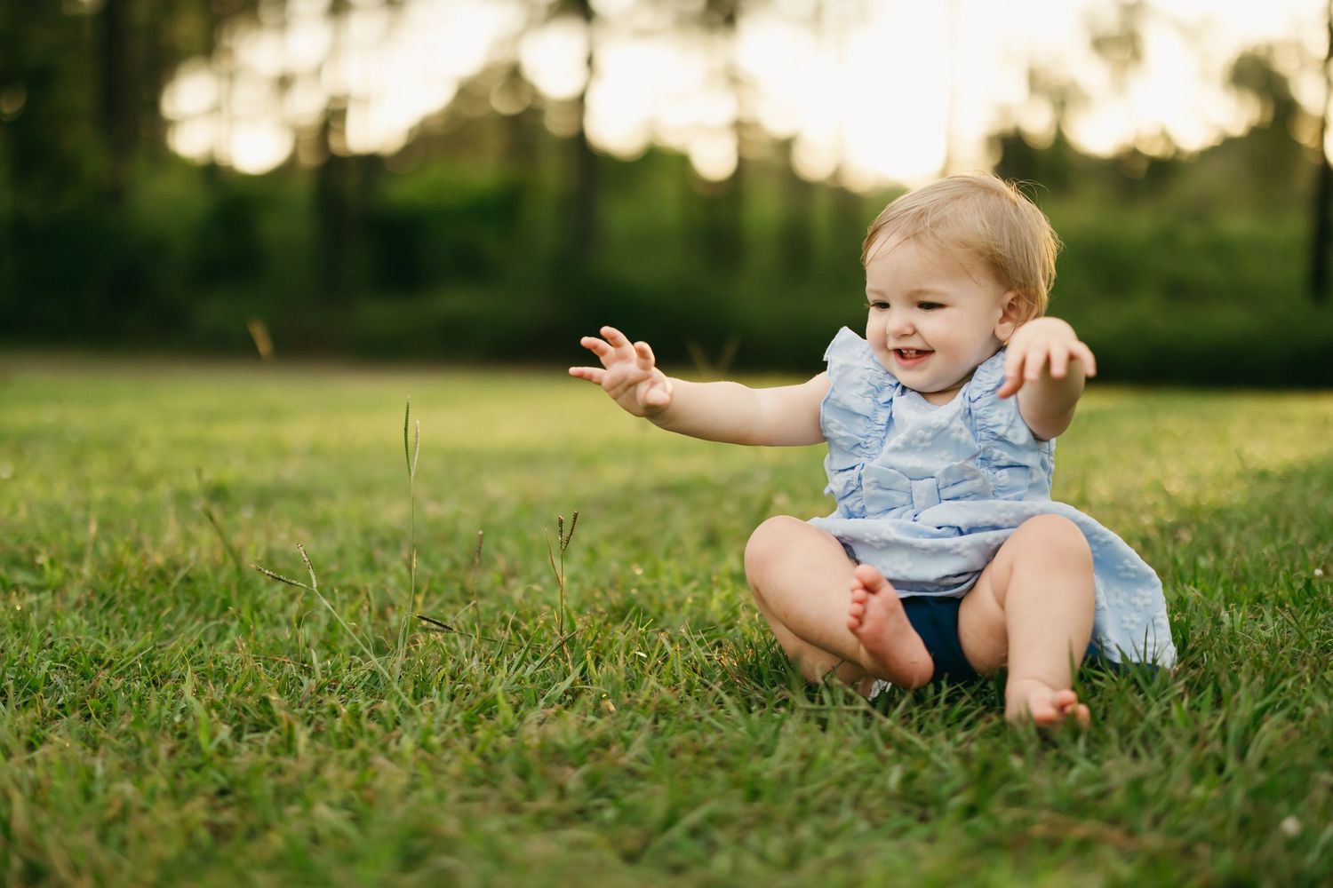 Baby wearing blue romper sitting and playing in grass during golden hour sunset.