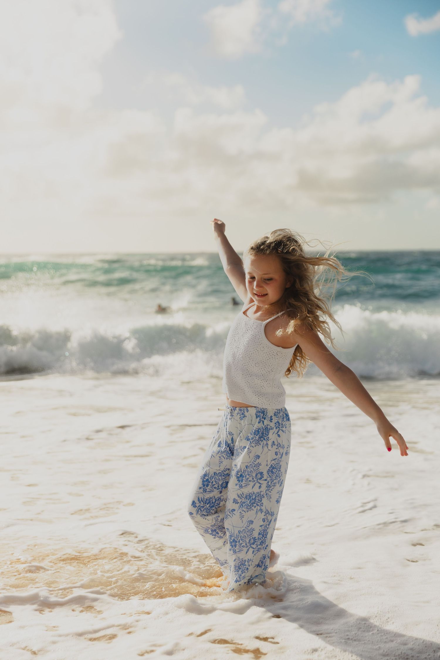 Figure in white dress and blue floral skirt dancing joyfully in ocean waves at beach.