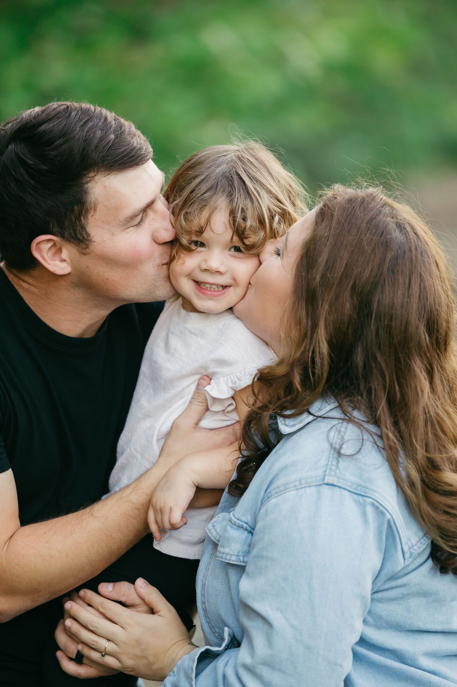 A loving family moment captured outdoors with parents kissing their young child's cheeks against a green background.