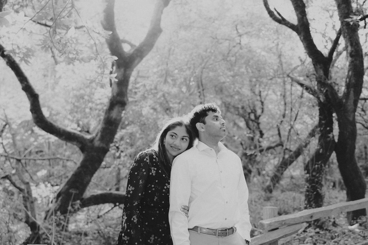 Black and white photo of a couple standing together among trees, woman leaning on mans shoulder outdoors.