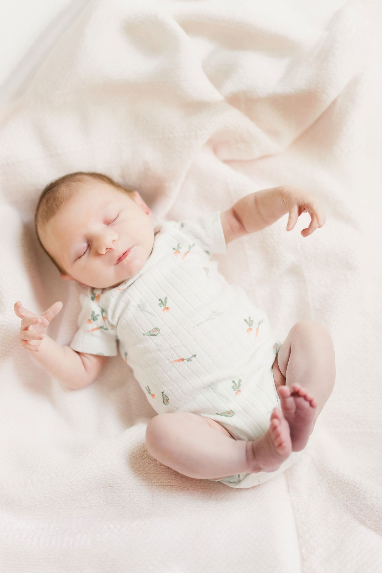 A sleeping newborn baby wearing a floral onesie lies peacefully on soft white bedding in a dreamy natural light setting.