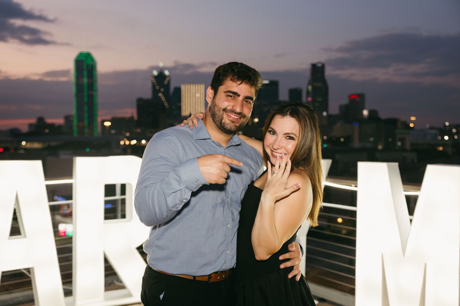 A romantic rooftop proposal scene at sunset with large white letters spelling MARRY ME against a city skyline.