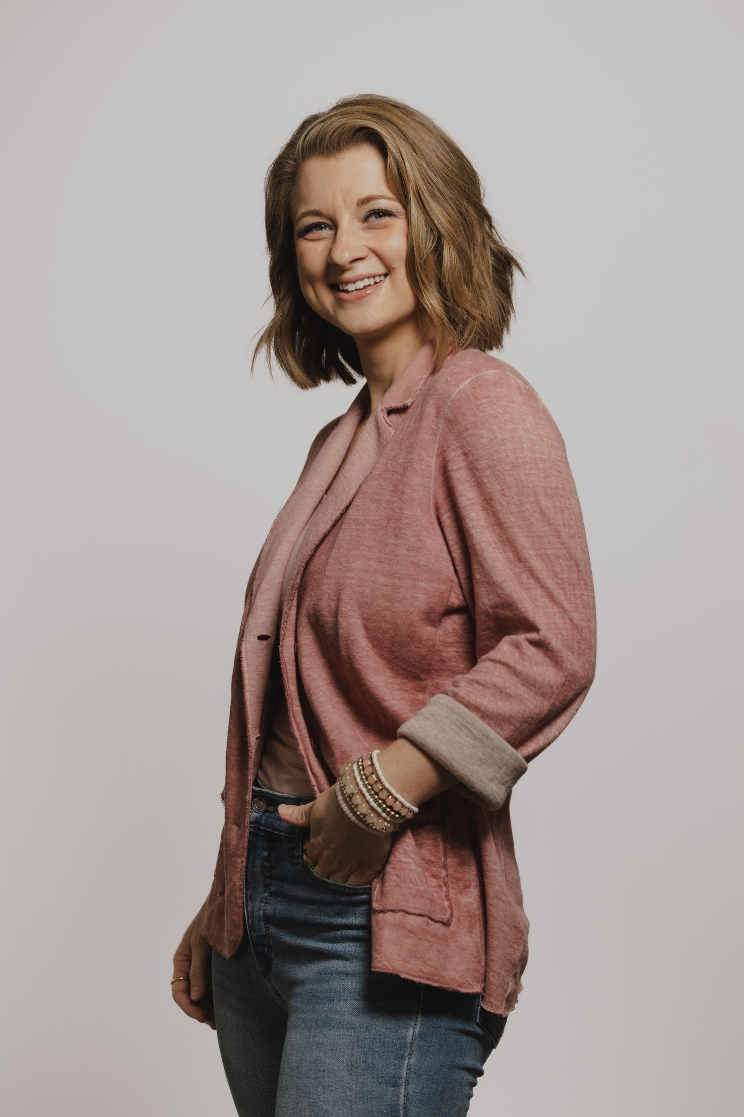 Professional studio portraits in a pink sweater and jeans against a gray backdrop.