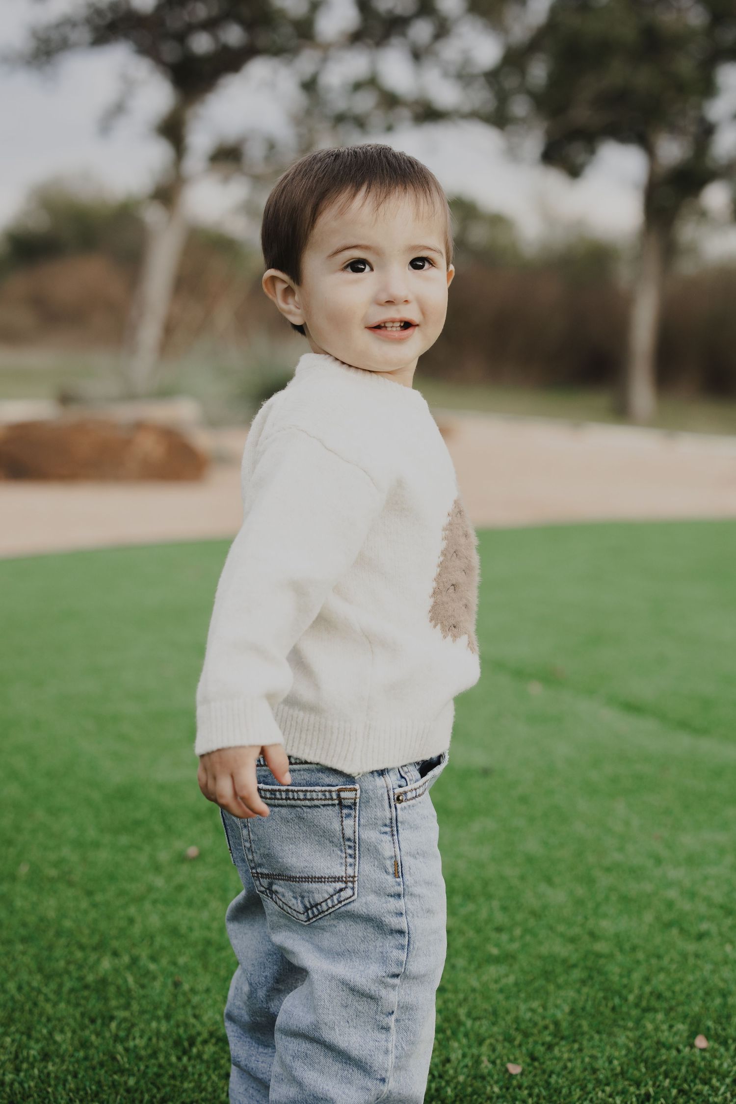 Small toddler wearing a white sweater and jeans standing on grass in an outdoor setting.
