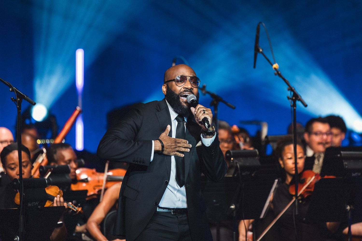 Male singer performing on stage in a black suit with an orchestra behind him under blue stage lighting.