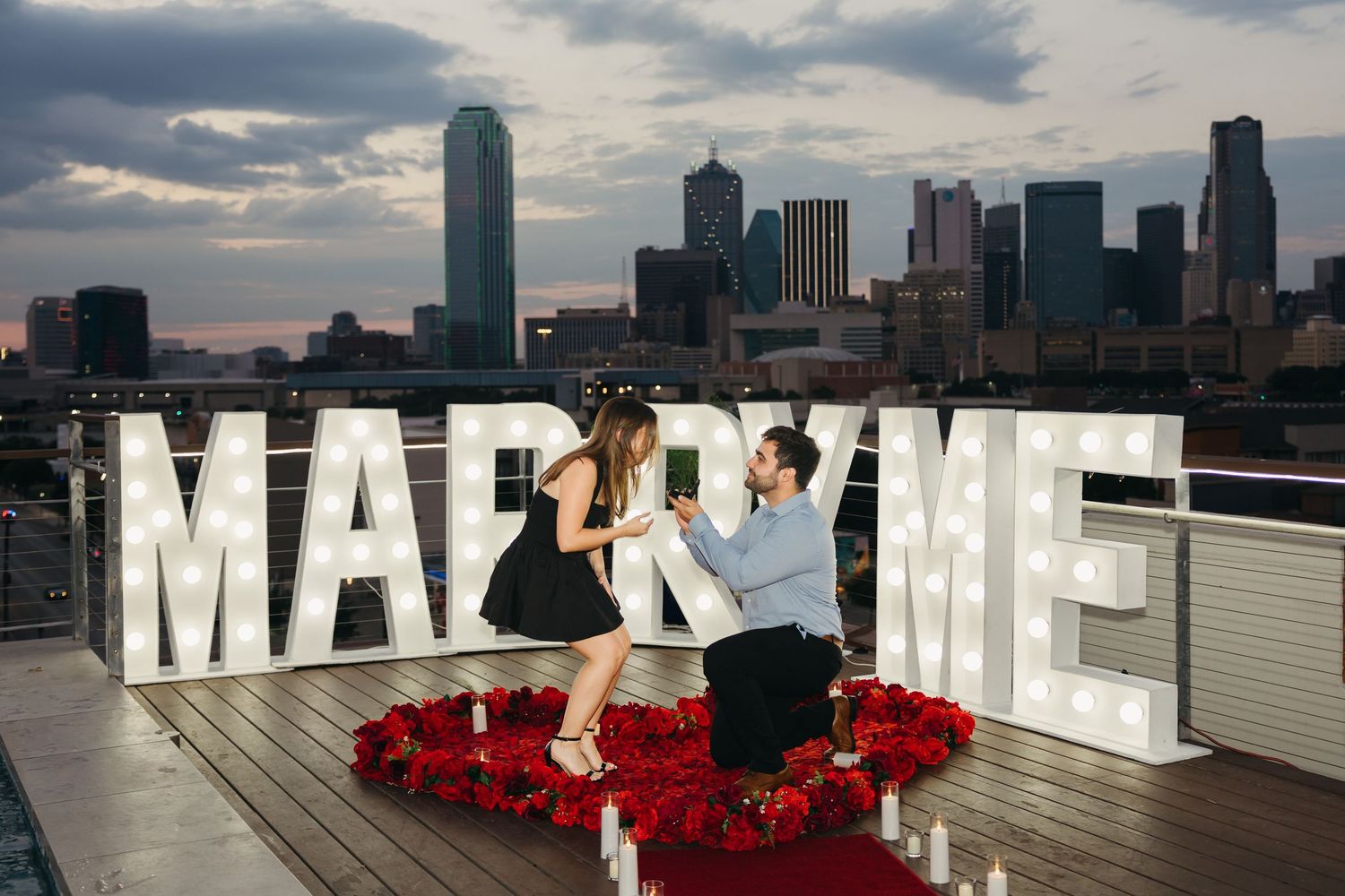 Rooftop proposal setup with large lit-up letters spelling 'Marry Me' against urban skyline.