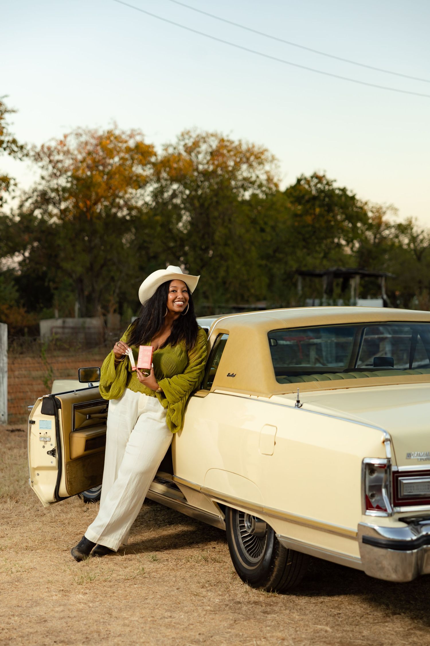 Woman in white hat and green blouse leaning against a vintage cream convertible car outdoors.