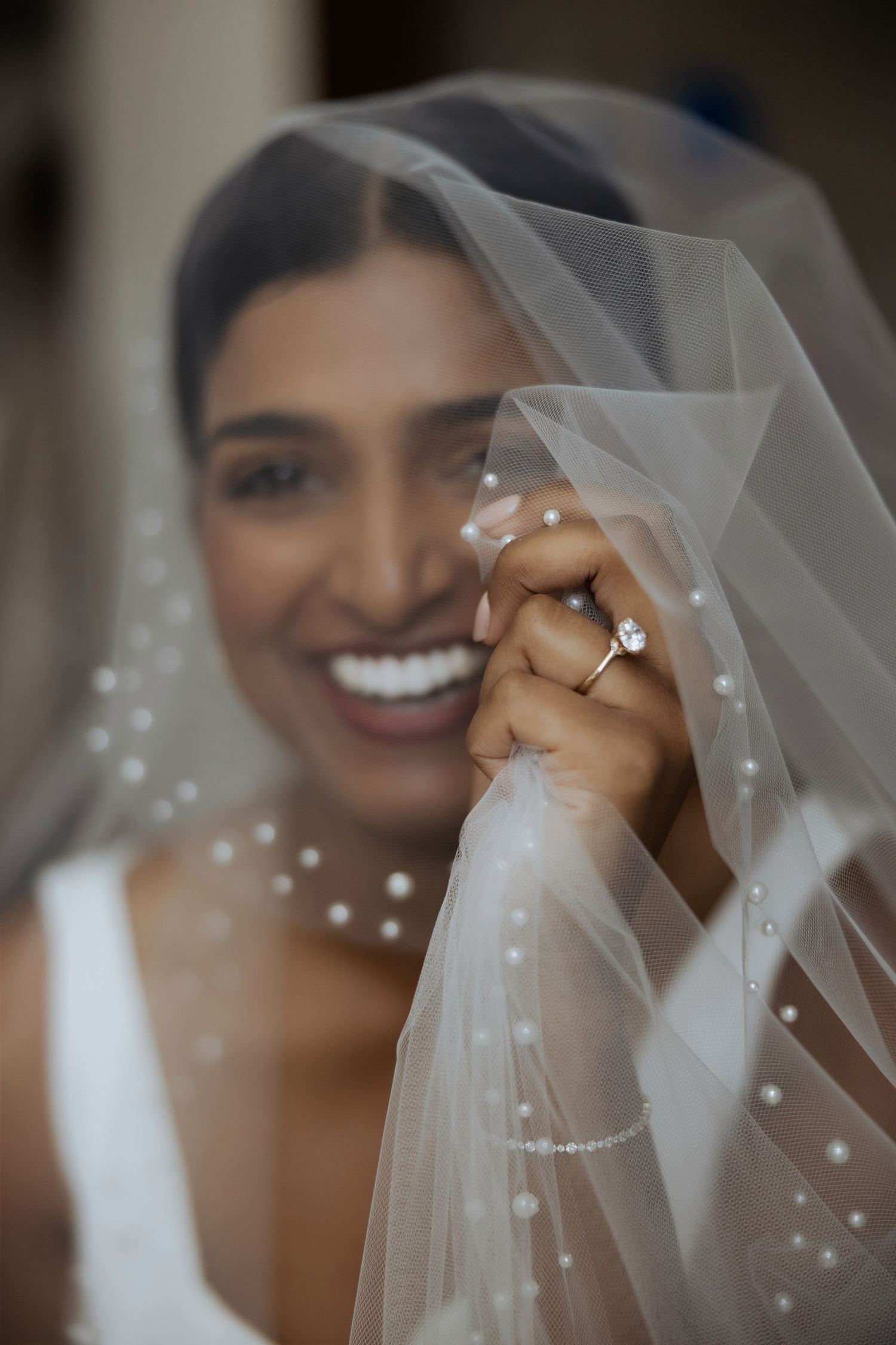 Smiling bride in white gown holds pearl-trimmed veil over face, showcasing diamond engagement ring.