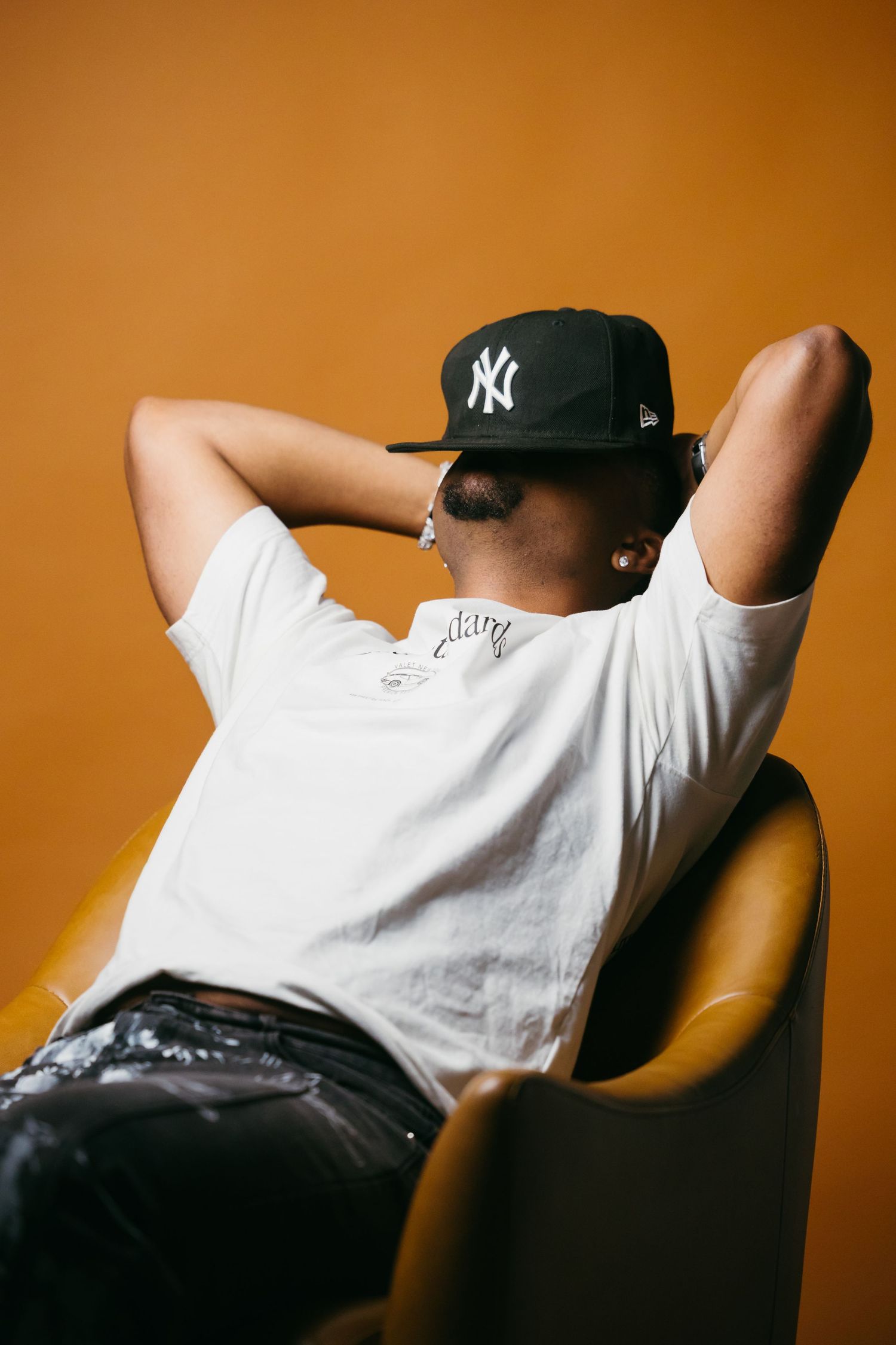 Person in white t-shirt and NY Yankees cap relaxes in chair against orange wall in artistic photo series.
