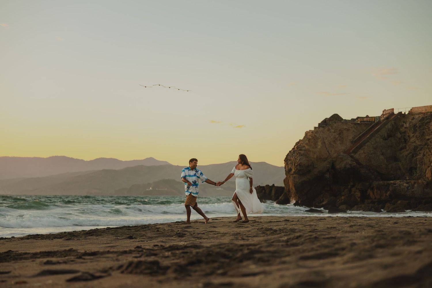 Couple holding hands walking on a sandy beach at sunset with ocean waves and rocky cliffs in the background.
